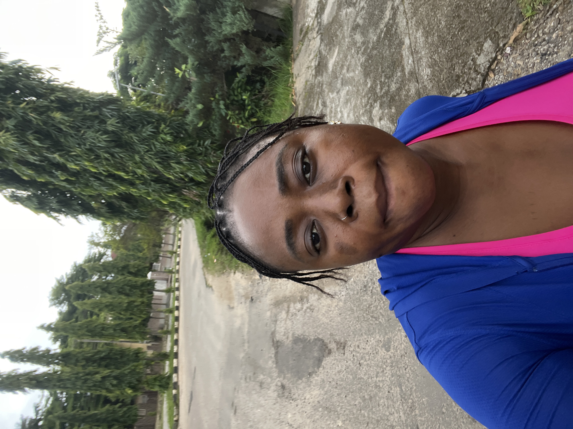 A medium-shot, eye-level selfie shows a Black woman with braided hair and a nose ring, looking at the camera with a neutral expression. She is wearing a blue zip-up jacket over a pink sports bra. The background reveals an outdoor scene in Abuja, Nigeria, with a paved road and sidewalk bordered by lush green trees and vegetation. The weather appears overcast.