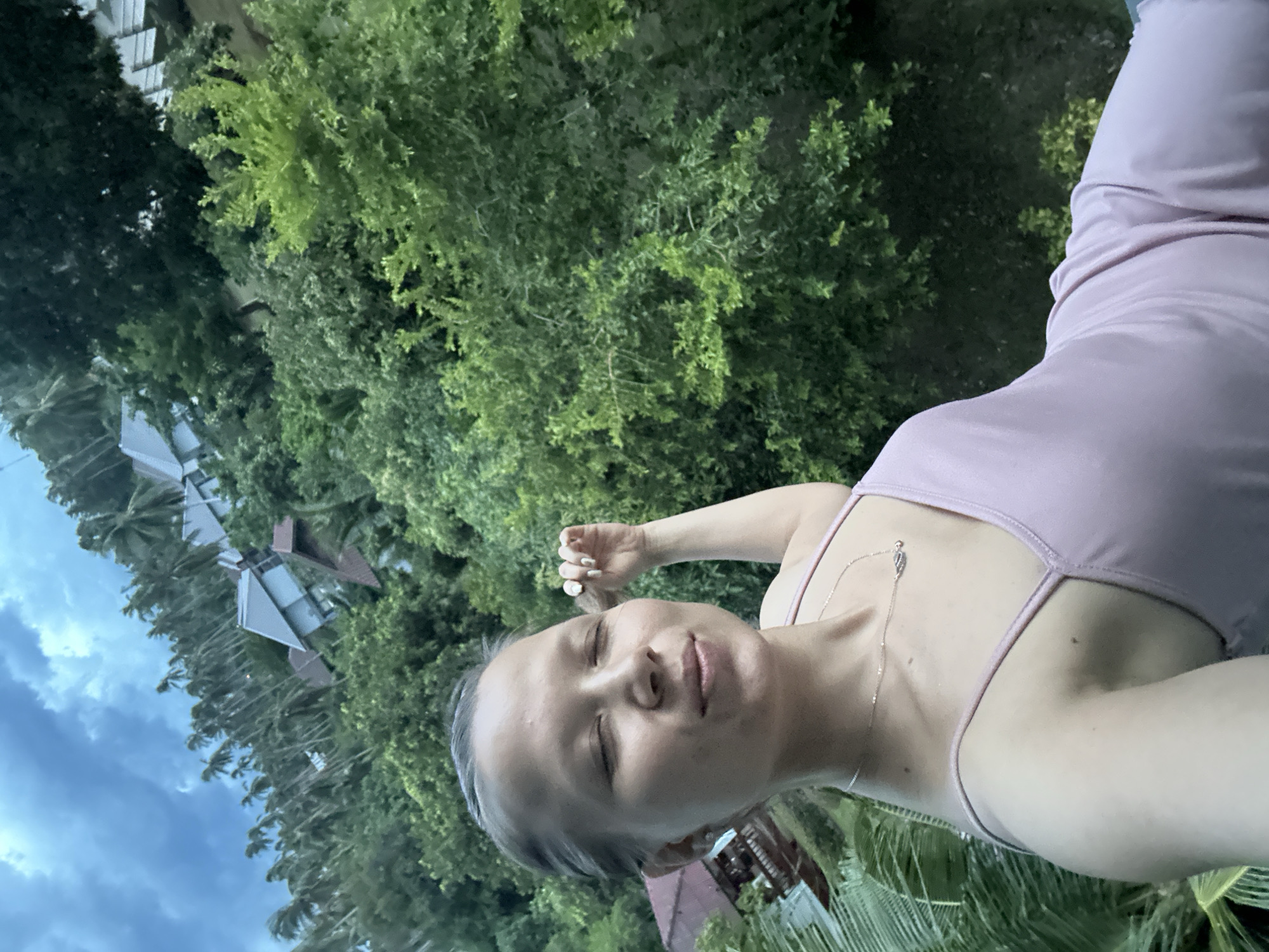 The image shows a young woman with her eyes closed, resting her head against her hand. She is wearing a light purple dress and a delicate silver necklace. The background is lush green foliage, with palm trees and tropical plants. In the distance, several modern buildings with triangular roofs are visible amongst the trees. The sky is overcast with dramatic clouds, suggesting it might be evening or a stormy day. The overall mood is peaceful and serene. The image appears to be a selfie, taken outdoors in a tropical location. The visible text is not legible.