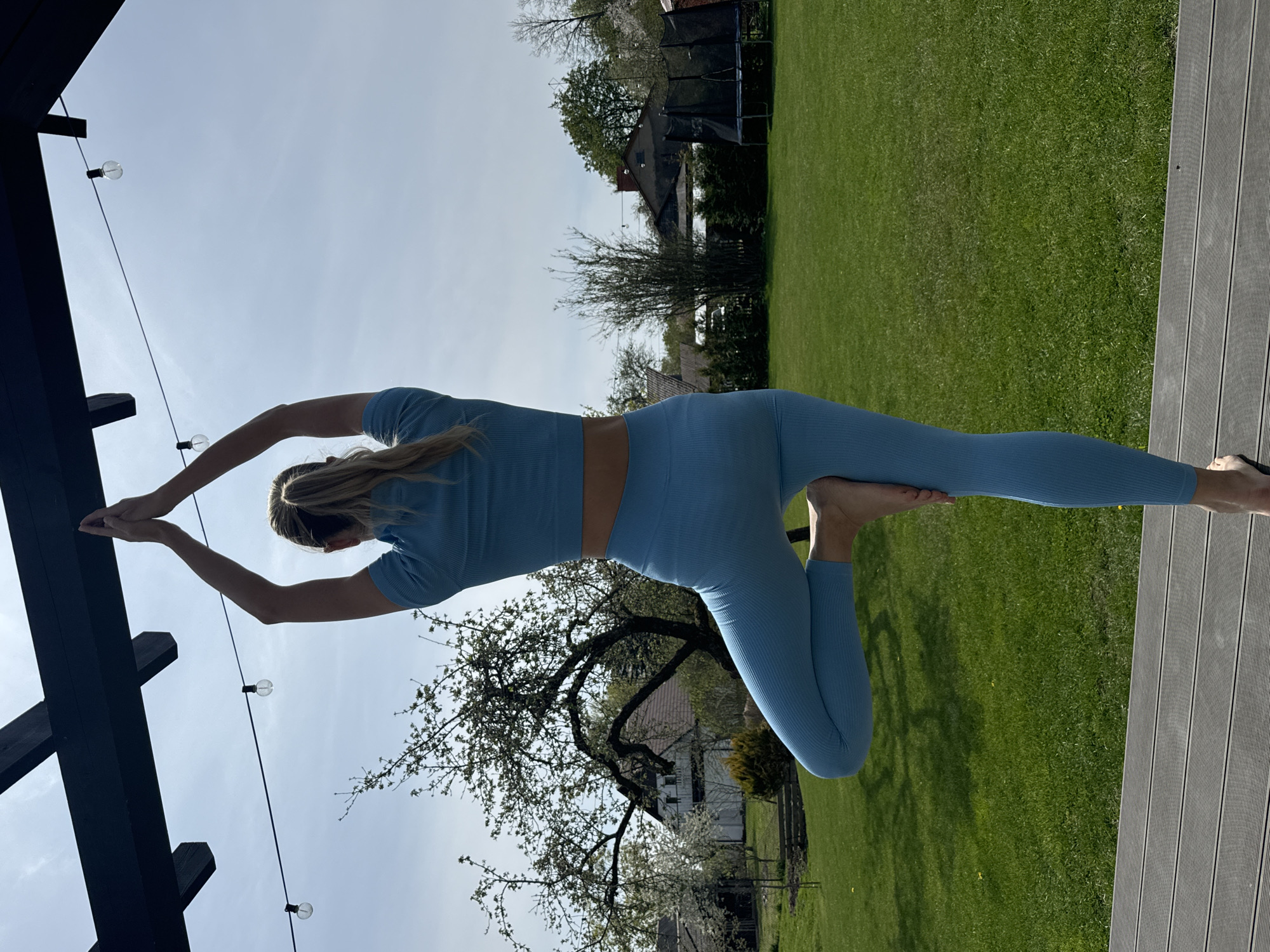A woman wearing a blue top and leggings is performing a yoga pose outdoors on a wooden deck. Her hands are placed on a dark wooden structure with string lights hanging from it. The background includes a green lawn, trees, a house, and a trampoline. The location is likely Carnikava, Latvia.