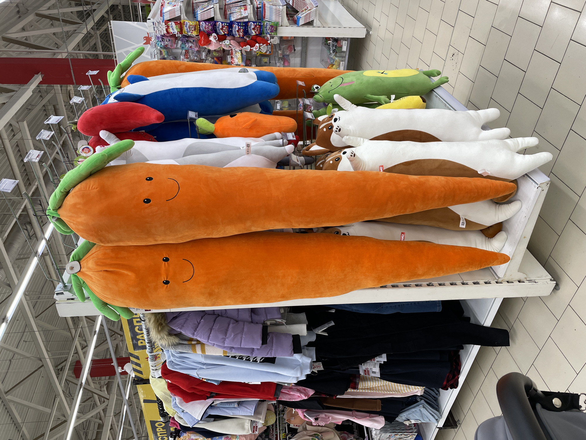 A vertically oriented image shows shelves in a store in Izhevsk, Russia. The shelves contain various items, including large, plush orange carrots with smiling faces, as well as plush figures shaped like a blue whale, a red whale, a green apple, and white and brown dogs. Below the top shelves is a shelf with clothing, including shirts and jackets. The store has tiled floors and bright overhead lighting. A stroller handle is visible in the bottom right corner.