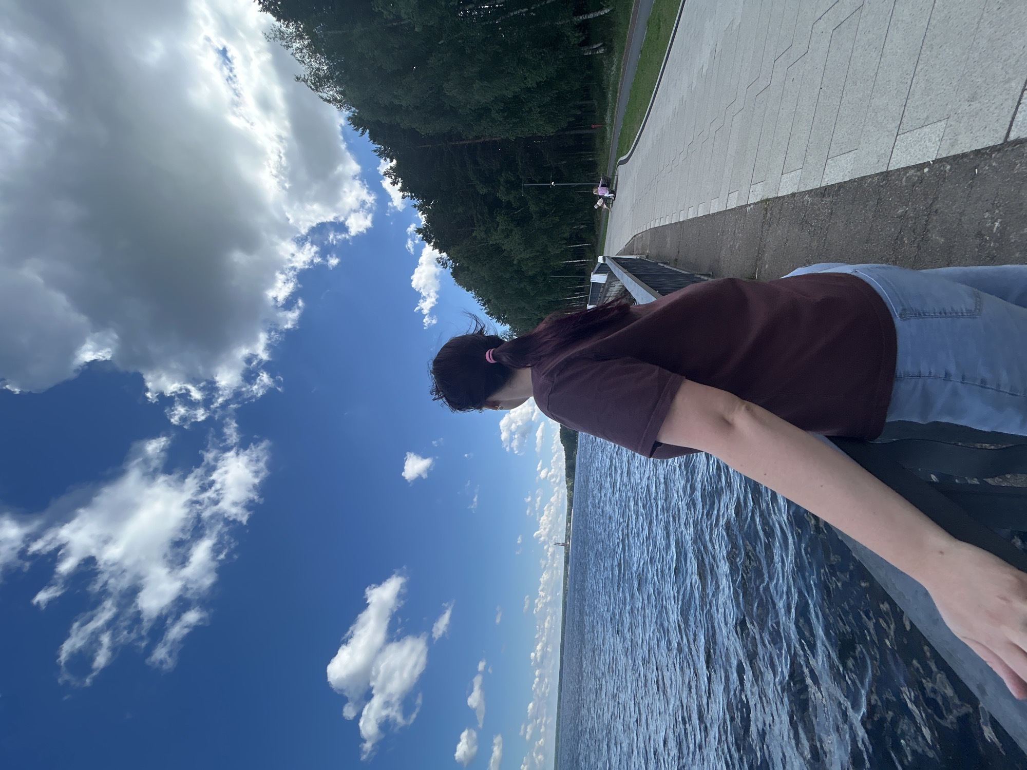 An over-the-shoulder shot shows a woman with dark hair pulled back in a ponytail, wearing a maroon t-shirt and light blue jeans. She is standing on a paved walkway next to a dark body of water. A railing separates the walkway from the water. In the distance, across the water, a red and white structure, possibly a tower, is visible against a bright blue sky dotted with white clouds. A dense line of green trees borders the walkway on the right side. Further down the walkway, two people are seated on a bench. The scene appears to be a promenade or embankment in the city of Chaikovsky, Russia, likely along the Kama River.