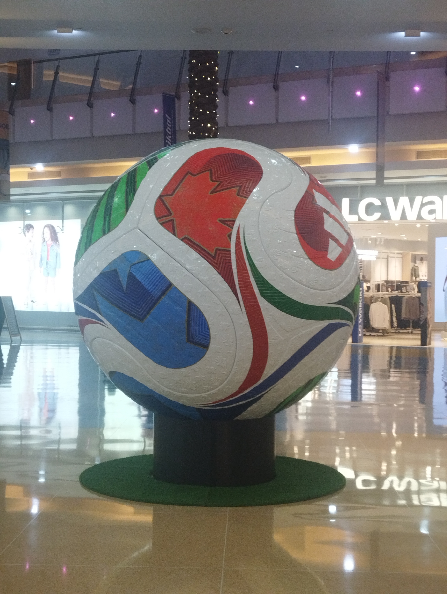 The image displays a large, colorful football (soccer ball) serving as a prominent centerpiece in what appears to be a modern shopping mall. The giant ball, designed with intricate patterns in white, red, blue, and green, resembles the Adidas Brazuca, the official match ball of the 2014 FIFA World Cup. It is mounted on a black cylindrical stand, which rests on a circular green mat, simulating turf.
The setting is indoors, characterized by a highly polished, light-colored floor that creates strong reflections of the ball, surrounding stores, and overhead lights. In the background to the left, a large illuminated display screen features two human figures, likely models for a clothing store. To the right, a retail establishment is visible with a prominent "LC Wal" sign, indicating it is an LC Waikiki store. Racks of clothing can be seen inside this store.
Above, portions of the mall's upper levels or balconies are visible, featuring glass railings and ambient lighting. Several square panels on the walls exhibit small, glowing pink or purple lights. Banners hang from the ceiling; one on the right is partially visible with the text "ikbal," and another on the far left shows unreadable text starting with "HOR." The scene is brightly lit by artificial lights, making the time of day indeterminate. The absence of people directly interacting with the ball suggests it's a static display, possibly for decoration or promotion within the commercial space.