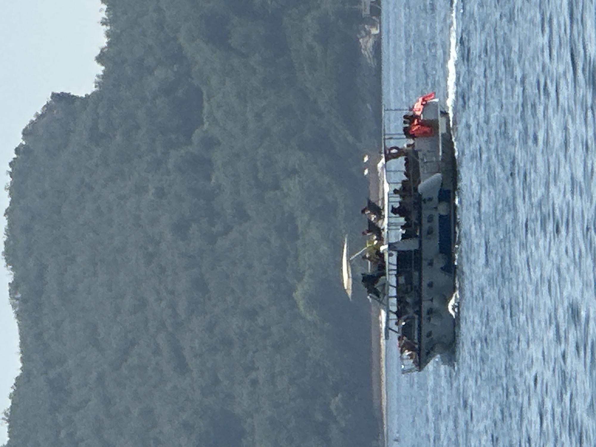 A tour boat filled with people is cruising through the blue waters of the Adriatic Sea, with the lush, green island of Sveti Stefan visible in the background. The boat appears to be a white passenger vessel with a covered upper deck and a white awning. Many individuals are standing and sitting on the deck, some wearing life vests in bright orange. The water is calm, with small waves rippling on the surface. The sky is a light blue, and the overall scene suggests a sunny day, likely during tourist season in Montenegro.