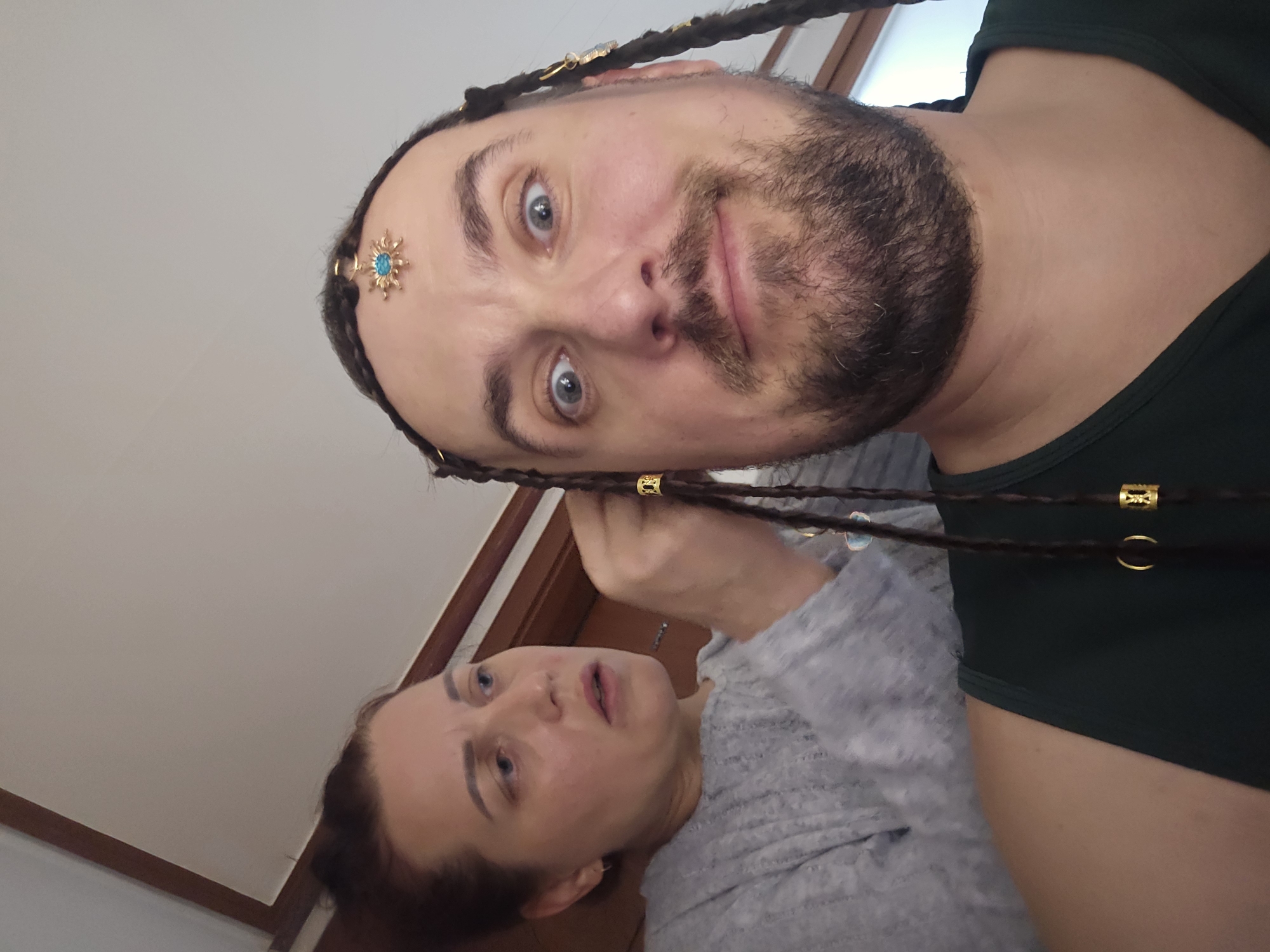 In a close-up, indoor, selfie-style photograph, a man with wide eyes and a beard has his hair being styled into braids by a woman behind him. The man is wearing a dark green tank top and a golden hair accessory with a blue gem is affixed to his forehead. The woman, wearing a grey sweater, is focused on her task, her mouth slightly agape. Several golden beads are visible in the man's braids. The background appears to be a domestic setting with a wooden trim around a ceiling. The image does not provide any specific visual cues to determine its location within Pyeongtaek-si, South Korea.