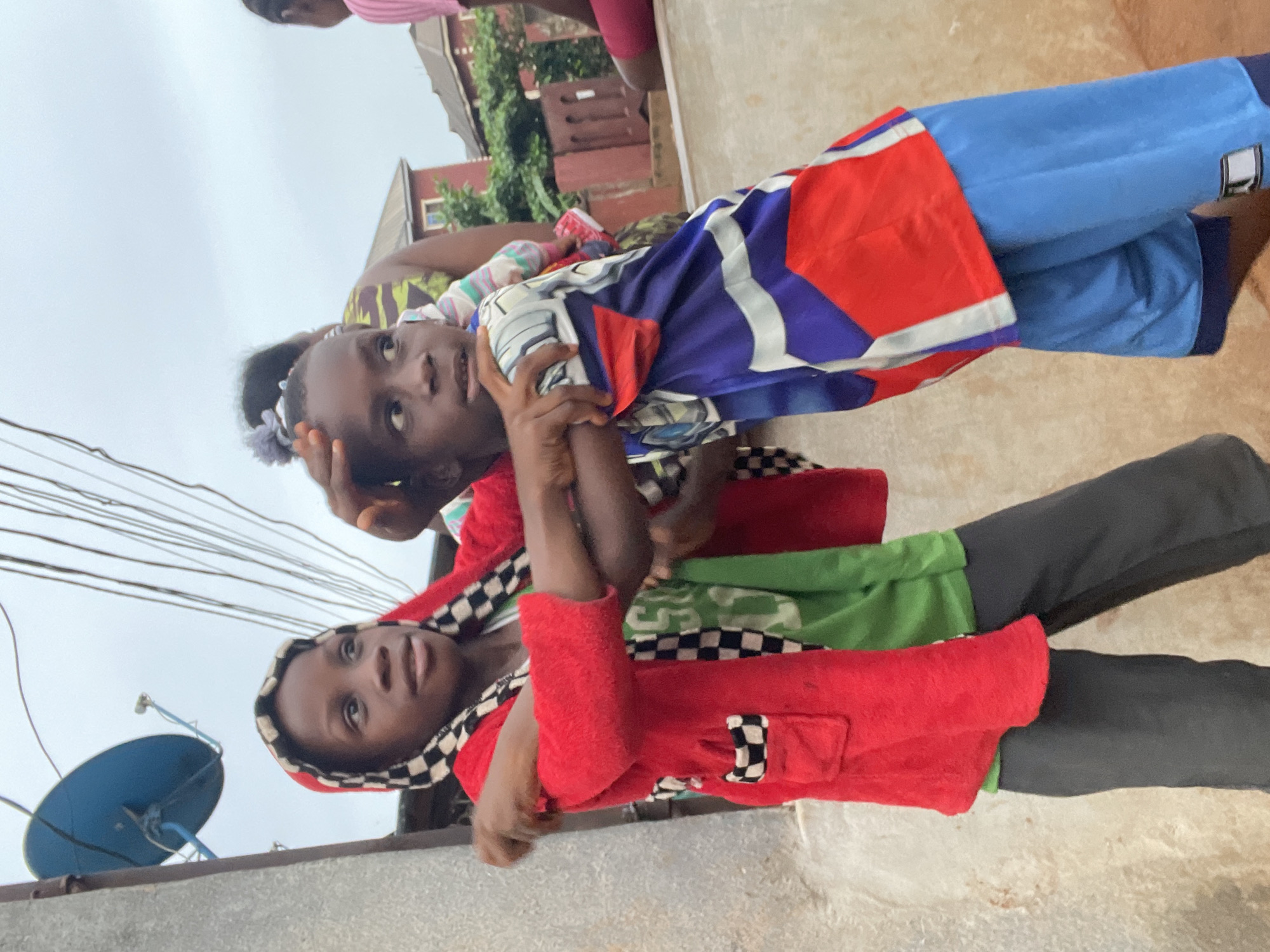 Two girls are posing on a rooftop in Onitsha, Nigeria. The girl closest to the camera wears a red hooded jacket with checkered trim, a green garment underneath, and gray pants. She looks directly at the camera. The other girl wears a white and gray headband, a patterned blue and purple top, a red jacket, and blue pants. A blue satellite dish is visible in the background. Overhead electrical wires are also present. A building and trees are visible further in the background.