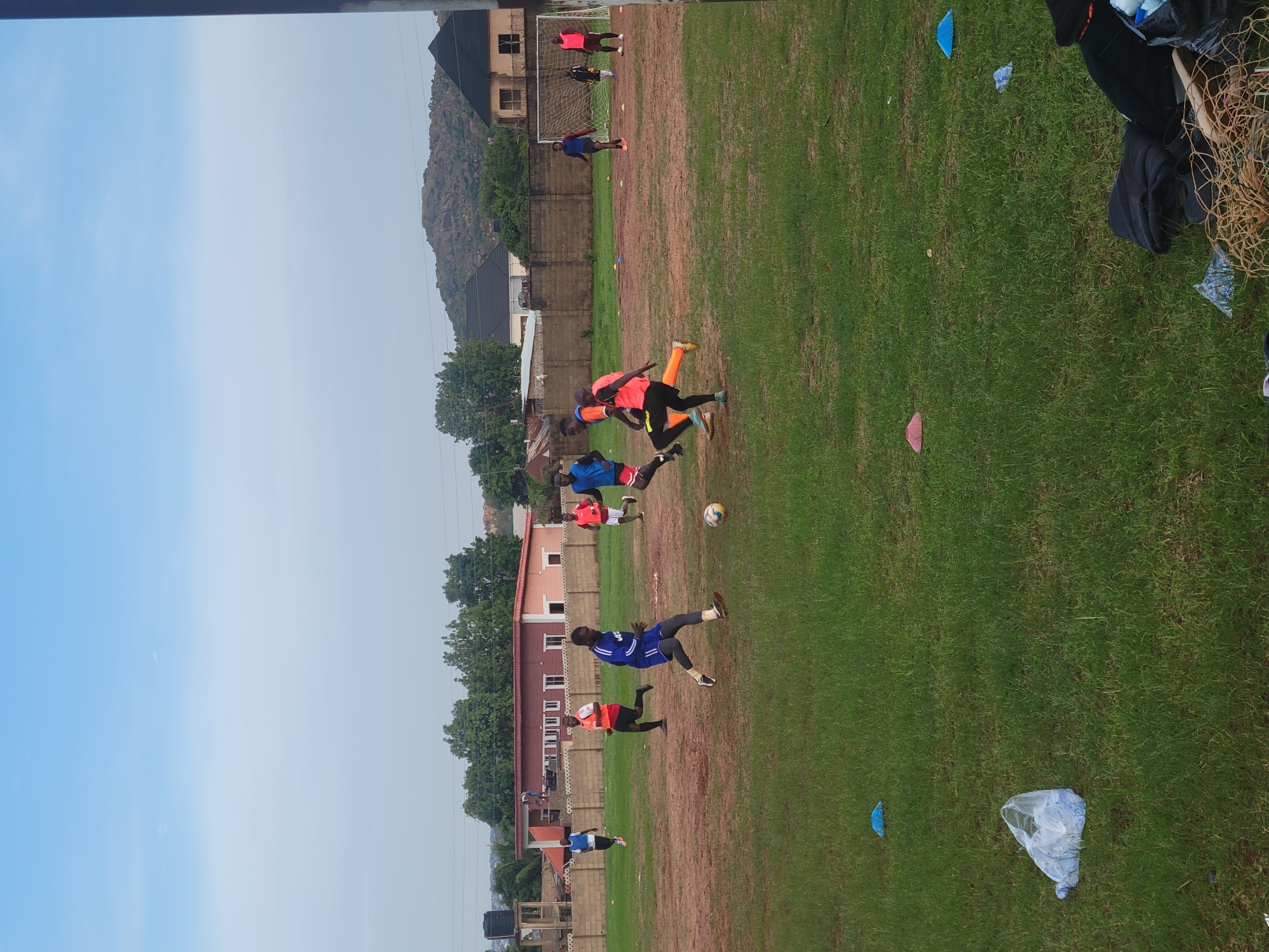 An outdoor scene captures a dynamic soccer match or training session taking place on a mixed grass and dirt field in Jos, Nigeria. Several individuals, primarily young men, are actively engaged in play.
In the foreground and mid-ground, players are scattered across the field. Two players in orange and black kits are central to the action, one appearing to be in motion to kick or control the ball, while another runs nearby. Slightly further back, a player in a blue kit is captured mid-air, seemingly jumping for a soccer ball that is visible just below him. Other players in blue, red, and orange jerseys are also seen moving across the pitch, with one person standing near the soccer goal in the background.
The playing surface consists of patches of vibrant green grass interspersed with areas of reddish-brown dirt. Small training cones, blue and pink, are visible on the field. Along the right edge of the frame, near the sidelines, there are discarded items including a black bag or jacket, a section of netting, and various plastic bags.
The background features low-rise buildings, likely houses, some with terracotta-colored roofs, behind a concrete wall. A significant rocky hill or small mountain range dominates the distant background, characteristic of the Jos plateau. The sky above is clear and bright, ranging from light blue to a hazy grey, indicating daytime. Scattered green trees add to the natural setting. The overall impression is one of active sports engagement in a local outdoor environment. No visible text is present.