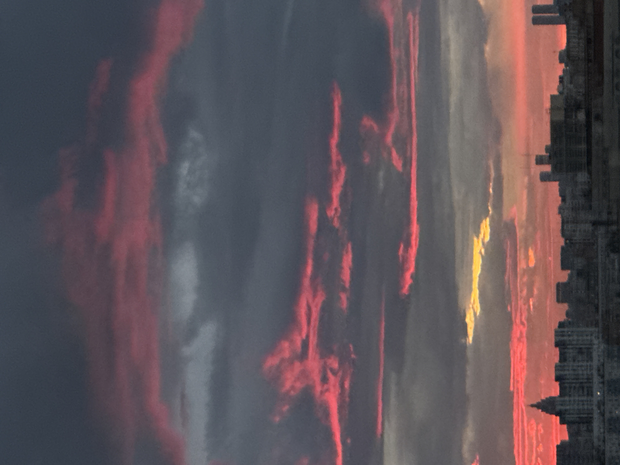 The image shows a cityscape at sunset in Moscow, Russia. The sky is predominantly gray with streaks of red clouds. A strip of yellow light is visible through the clouds. The silhouettes of buildings form a dark line on the right side of the image.
