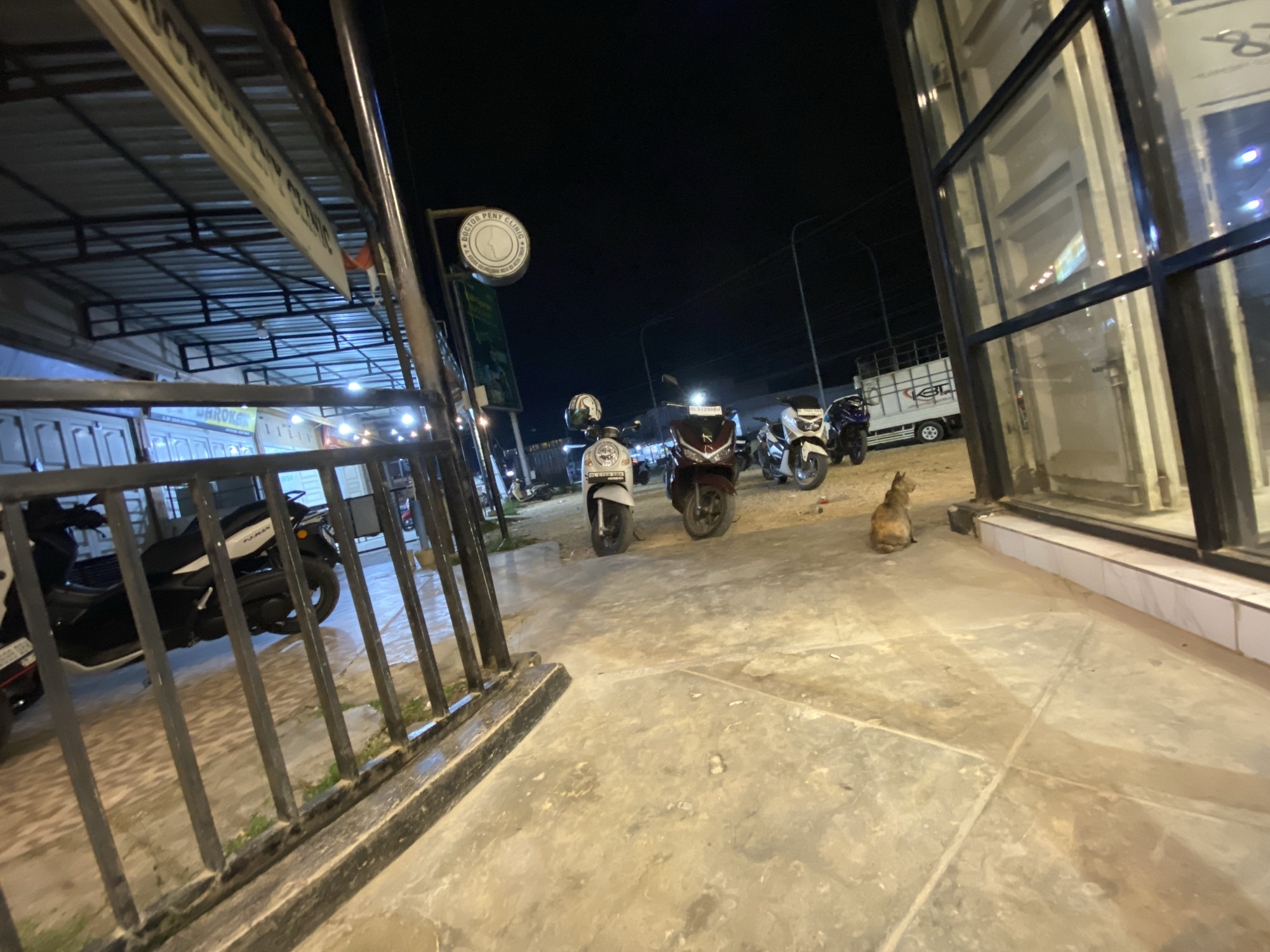 The image, taken at night in Keudeublang, Indonesia, depicts a street scene with several parked motorbikes and a solitary cat. In the foreground, a black metal fence partially obscures the view, behind which are parked motorbikes. Further into the scene, four additional motorbikes are visible, parked adjacent to the building on the right. A cat is seated next to this building. The building itself features large glass windows with black frames. Above the motorbikes, a sign reading "DOCTOR PENY CLINIC" is displayed. A truck is visible in the background, slightly to the right. The area is illuminated by streetlights and lights from the nearby buildings. The ground is paved with light-colored concrete slabs. The lighting conditions suggest it is nighttime.