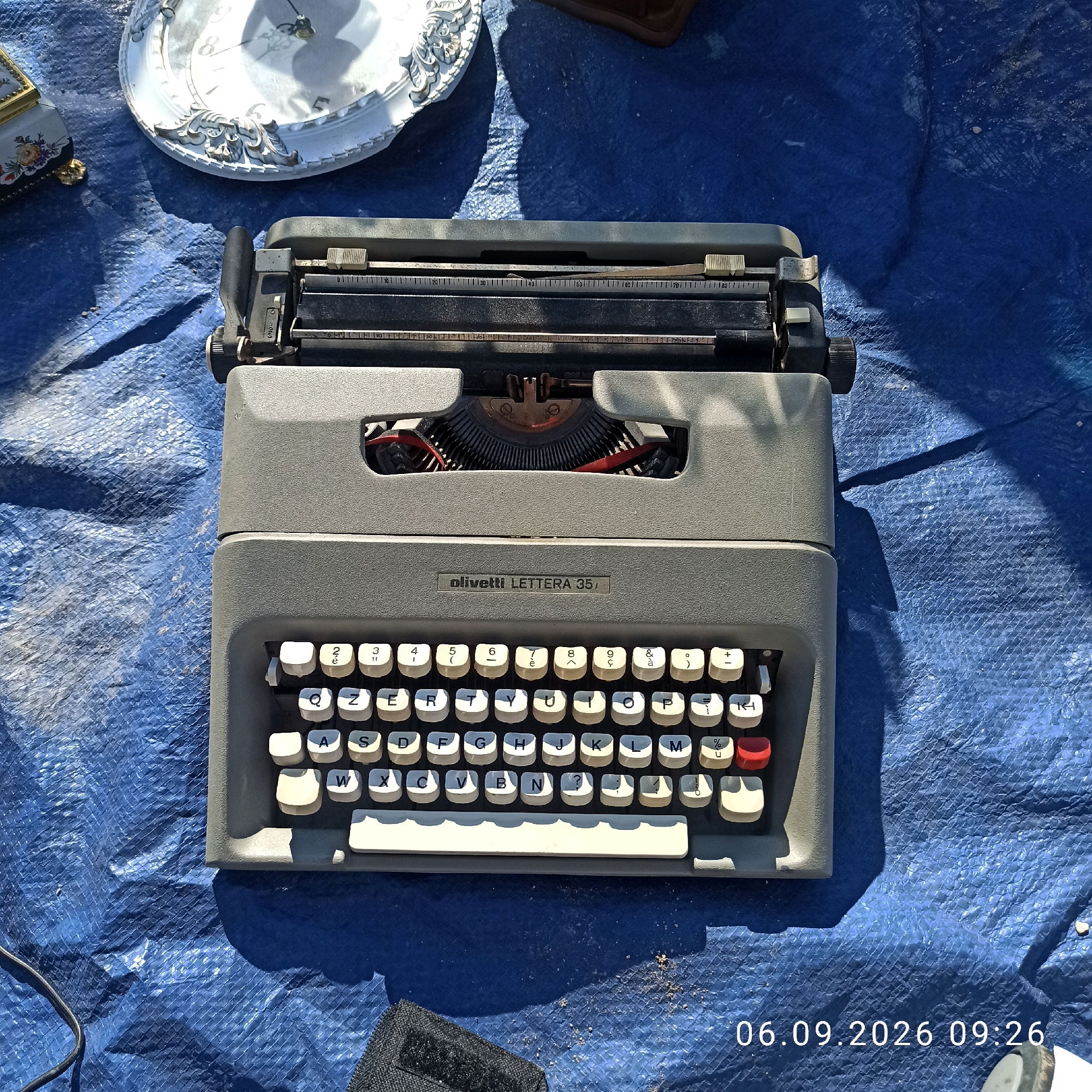 The image shows a vintage Olivetti Lettera 35 typewriter placed on a blue tarp. The typewriter is grey and has white keys with black lettering. The brand and model name "olivetti LETTERA 35" are visible on the front of the typewriter. In the background, a portion of a decorative white plate and a small ornate box are visible. The scene is outdoors, with dappled sunlight and shadows cast on the tarp. A black cable snakes across the lower left corner of the frame. The bottom right corner displays a date and time stamp: "06.09.2026 09:26". There are no people or visible interactions in the scene. The location is Sannicandro di Bari, Italy.