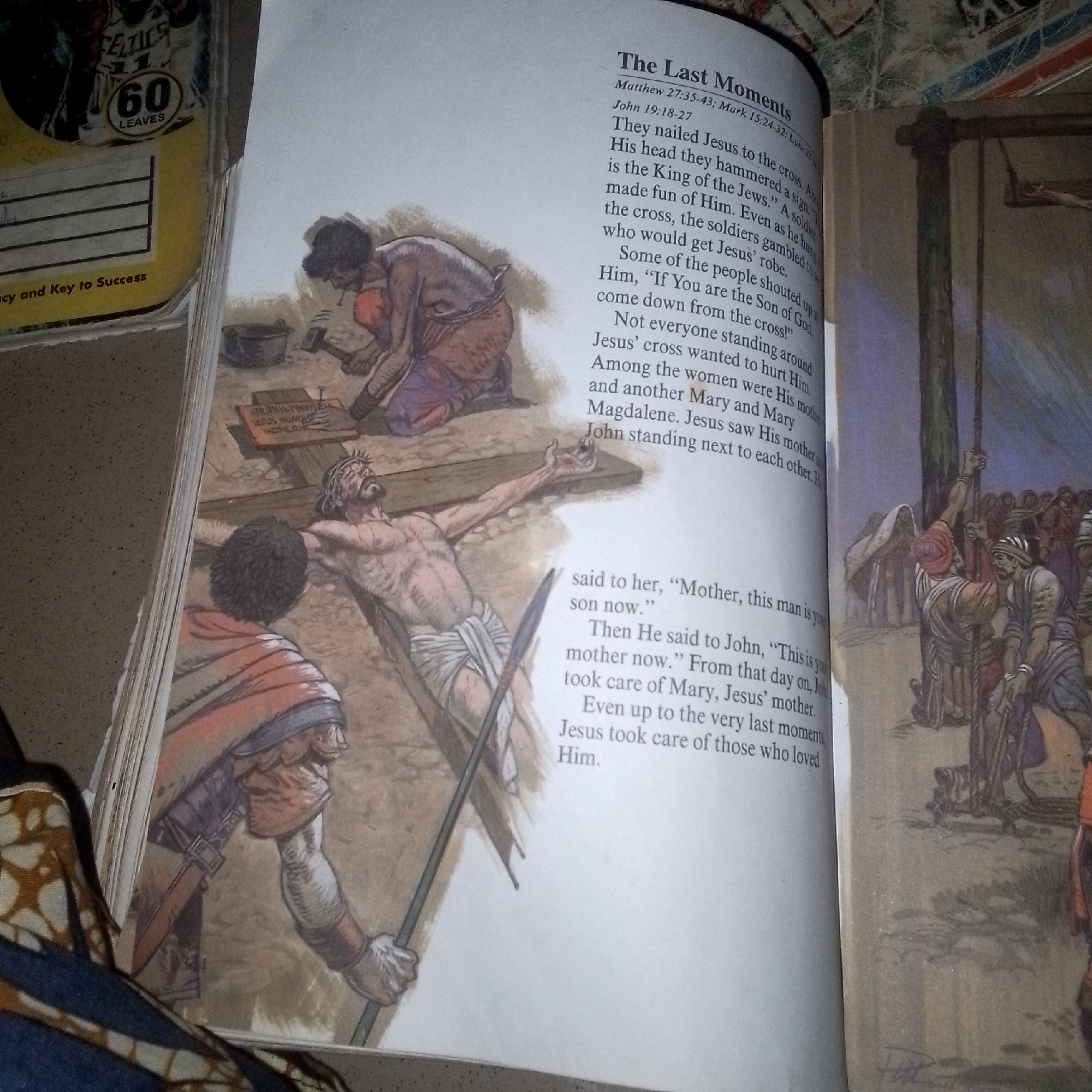 The image depicts a religious illustration with text. On the left, there is a partial view of a book or pamphlet with text and a graphic that appears to be a product label with "60 LEAVES" written on it. The main illustration shows a scene of the crucifixion of Jesus Christ. Jesus is depicted on the cross, with a crown of thorns on his head and a loincloth. Two figures are actively involved in the crucifixion: one is hammering a nail into Jesus' hand, and another is standing nearby, holding a spear. Another figure, possibly a Roman soldier, is seen in the background with a group of people. To the left of the crucifixion scene, there's another illustration of a person on their knees, using a hammer. The text on the right page is titled "The Last Moments" and appears to be a biblical passage, likely related to the crucifixion, with references to Matthew, Mark, and John. The style of the illustration is realistic with muted colors. The scene is set outdoors, on what appears to be a rocky or dusty ground.