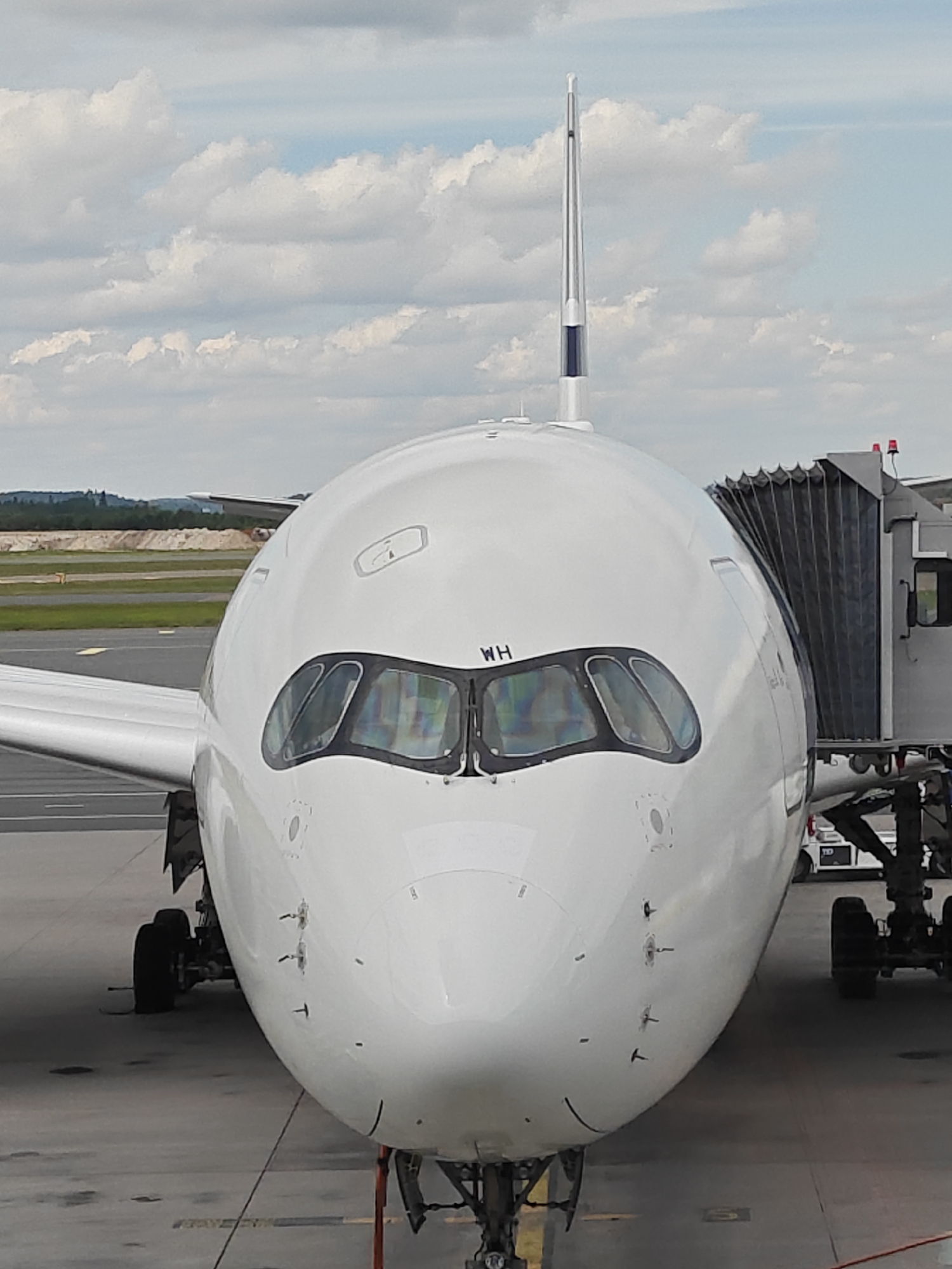 This image shows the front of a white airplane, possibly an Airbus A350, parked at a gate. The airplane is equipped with a jet bridge, indicating it is at an airport. The registration mark "WH" is visible on the nose of the aircraft, near the cockpit windows. The scene appears to be at Helsinki Airport in Vantaa, Finland, given the country-specific information. The sky is partly cloudy, and the ground is asphalt, typical of an airport tarmac. No people are visible in the image. The airplane is stationary, likely awaiting passengers or preparing for departure.