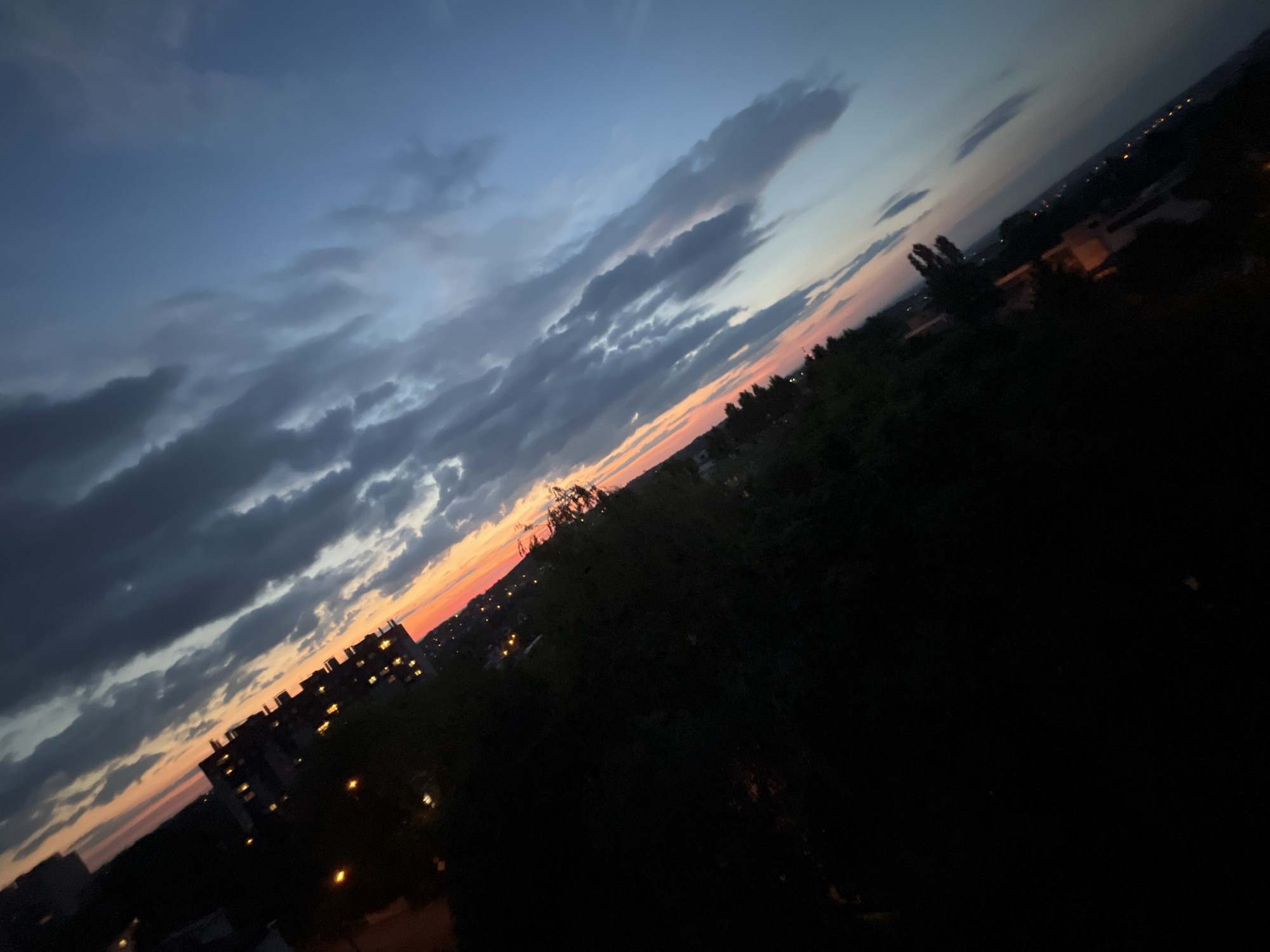 The image shows a tilted view of a cityscape at dusk. The sky is a gradient of deep blue at the top, transitioning to hues of orange and pink near the horizon, indicative of a sunset. Several dark, elongated clouds stretch across the sky diagonally. In the foreground and midground, silhouettes of trees and buildings are visible. One prominent building in the lower left quadrant is a multi-story structure with illuminated windows. Streetlights and other scattered lights suggest a populated area. The overall scene is dark, with the vibrant sky providing the main source of light and color, contrasted by the shadowy urban landscape.
