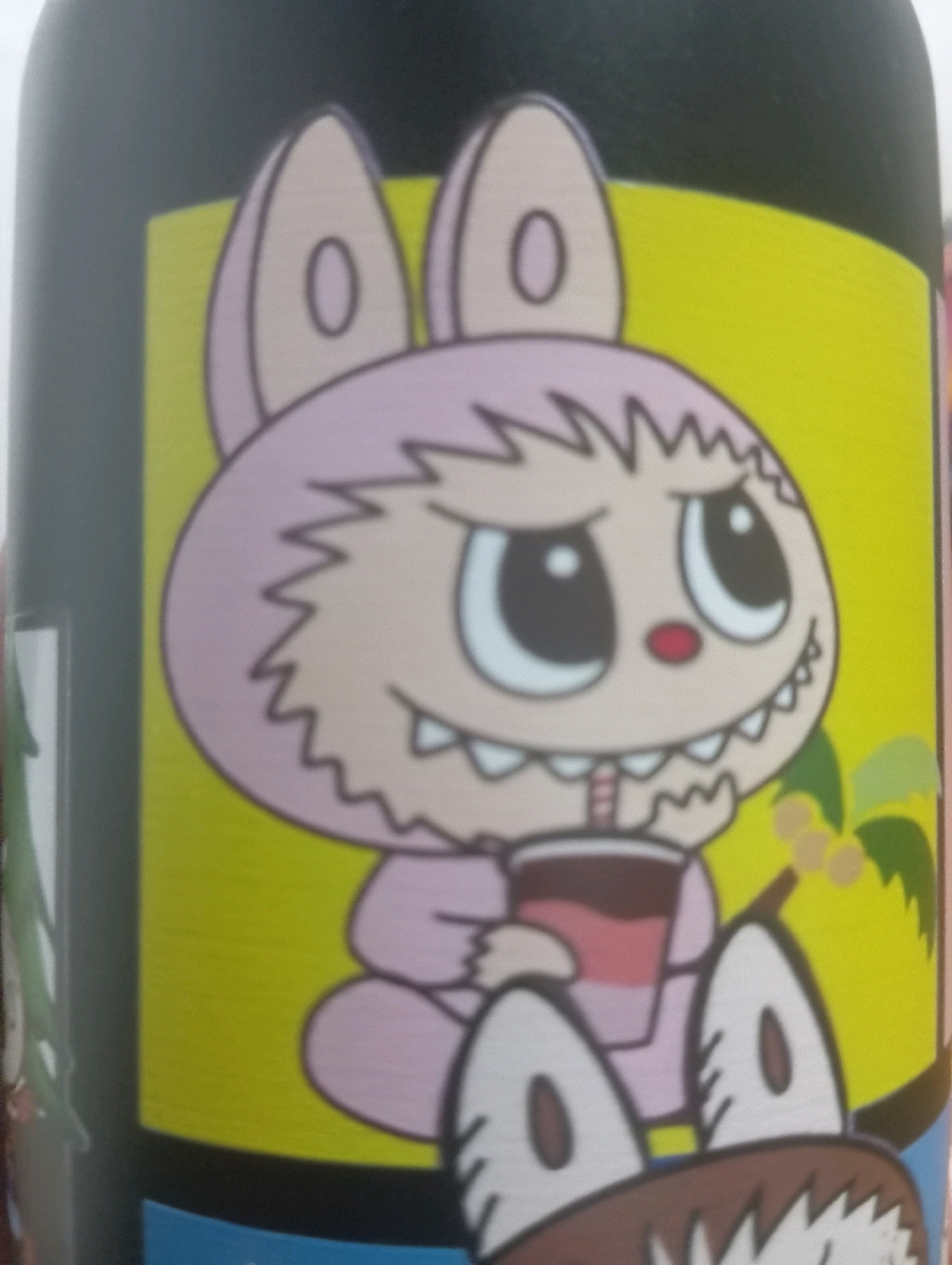 The image is a close-up of a cartoon drawing on a dark cylindrical object, likely a bottle or can. The drawing features a pink, rabbit-like character with large eyes and a wide mouth showing sharp teeth. The character is wearing a pink outfit and holding a straw in a dark beverage. To the right of the character, there is a depiction of a palm tree with yellow fruit. The background is a bright yellow. The object is positioned on what appears to be a dark surface, possibly a table or shelf. There is no discernible location within the city of Kutasari, Indonesia, as the image focuses solely on the illustration.