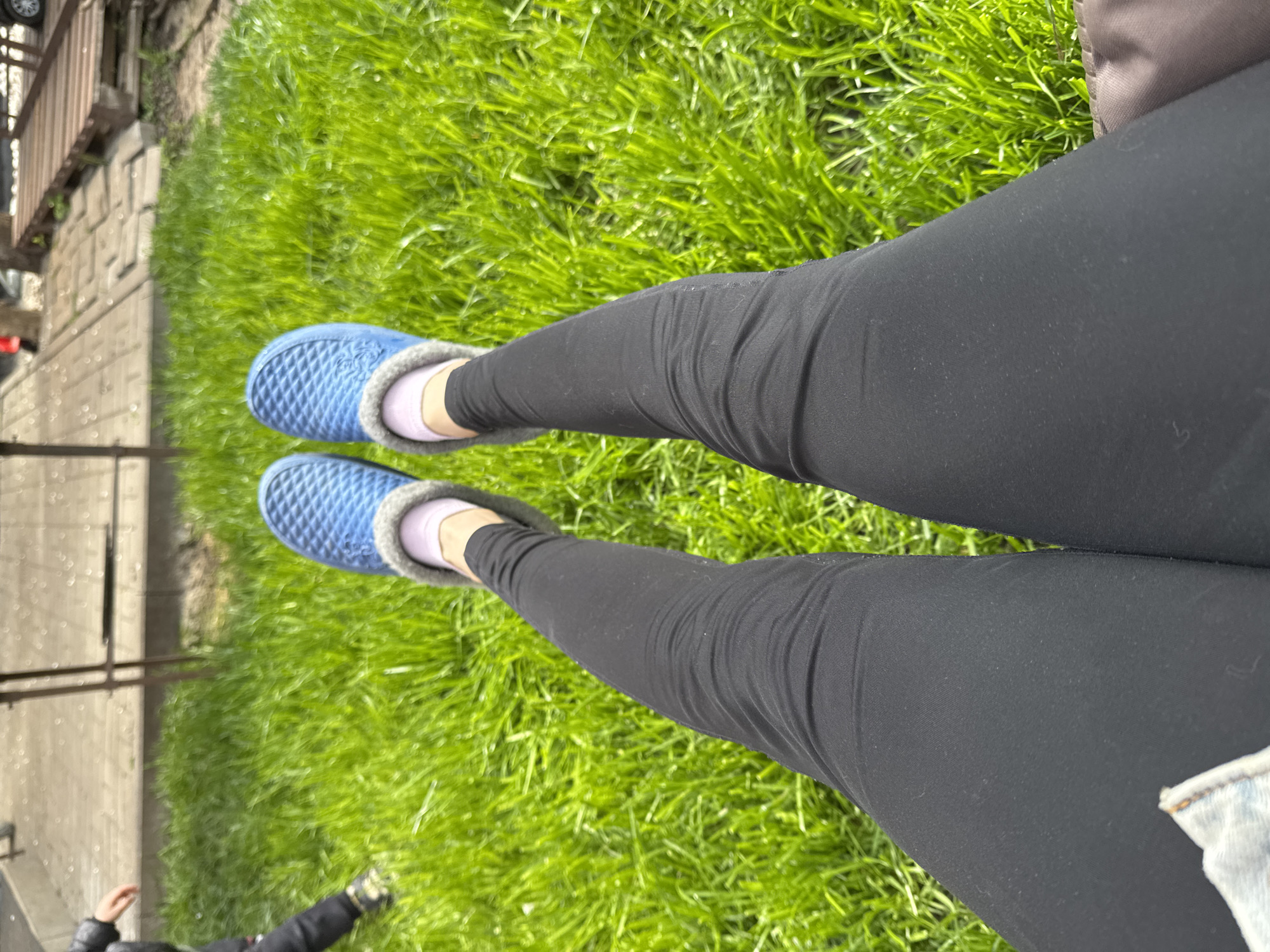 A high-angle shot shows a person's legs clad in black leggings, wearing blue quilted slippers over pink socks, resting on green grass. The setting appears to be outdoors, possibly a park or grassy area in the city of Rostov-on-Don, Russia, as suggested by the prompt. A concrete sidewalk or path is visible on the left side, along with a metal railing. A small portion of a denim jacket is visible near the lower right corner. The photo is taken in daylight.