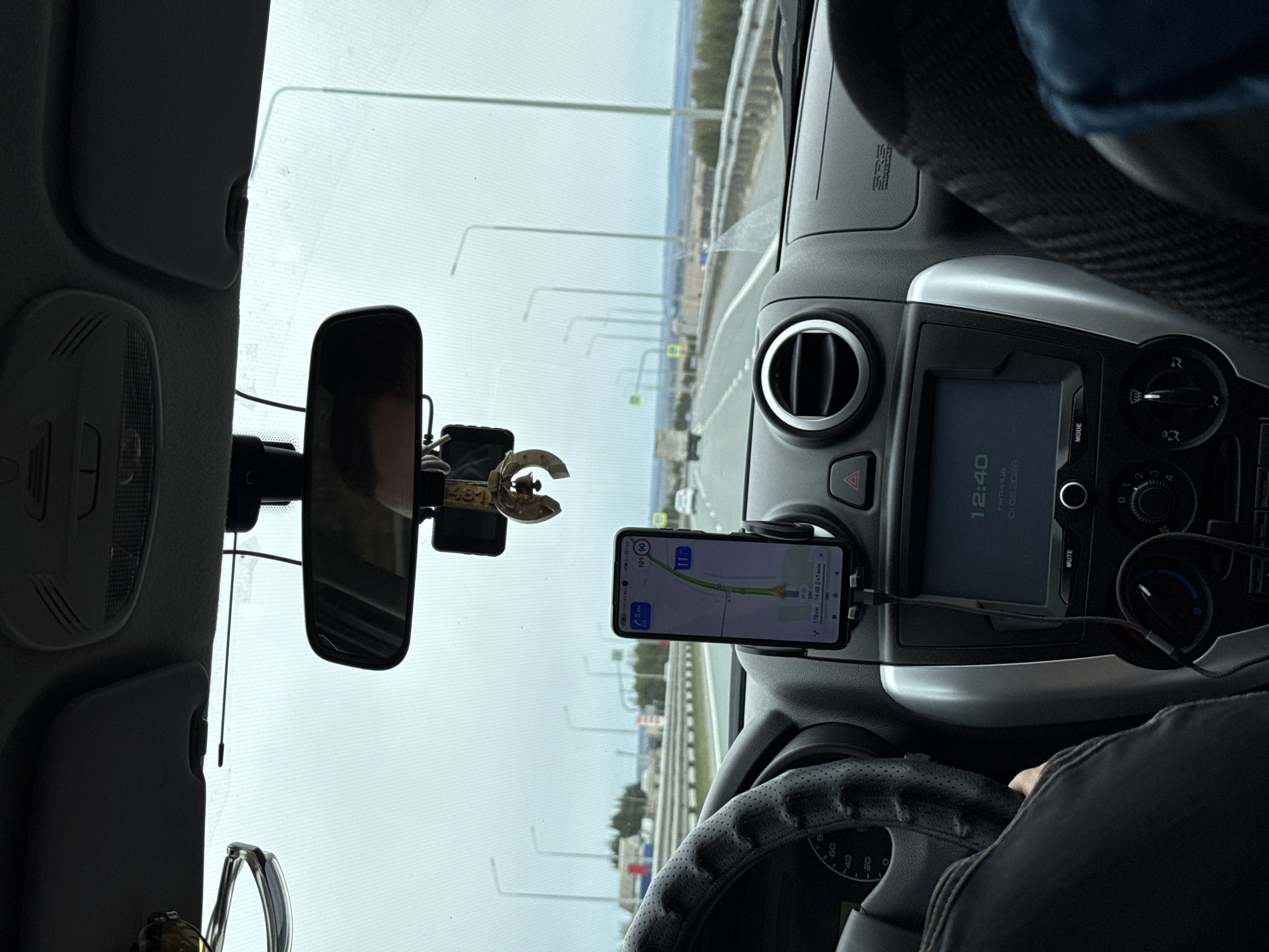 The image provides a driver's perspective from inside a car traveling on a multi-lane highway or wide road, likely in Покровское, Russia. The sky outside appears overcast and bright, indicating daytime.
The primary subjects include the car's interior and the road ahead. A driver's arm and leg are visible on the right, with their hand resting on a black, perforated steering wheel. A smartphone, mounted centrally on the dashboard, displays a navigation application showing a route. The navigation indicates "178 км" (178 km), an estimated arrival time of "14:40", and "2 ч 1 мин" (2 hours 1 minute) remaining. The phone's screen also shows a current speed of "50" (likely km/h) and a speed limit of "11".
On the car's built-in display, the time "12:40", the day "пятница" (Friday), and the date "01.05 2026" are visible. Below this, buttons for "MUTE" and "MODE" are present, along with climate control knobs. An "SRS AIRBAG" label is on the passenger side of the dashboard. A rearview mirror hangs in the center, reflecting a blurred image, possibly of the driver. A small, metallic charm, possibly a horseshoe shape, displaying "1487" or "14871" is attached near the rearview mirror.
The scene depicts a typical long-distance journey, with the driver using navigation for guidance. The presence of streetlights or gantries lining the road suggests a developed infrastructure. The time difference between the car's clock (12:40) and the navigation's estimated arrival time (14:40) is notable, possibly due to time zone settings or an incorrectly set car clock given the future date.