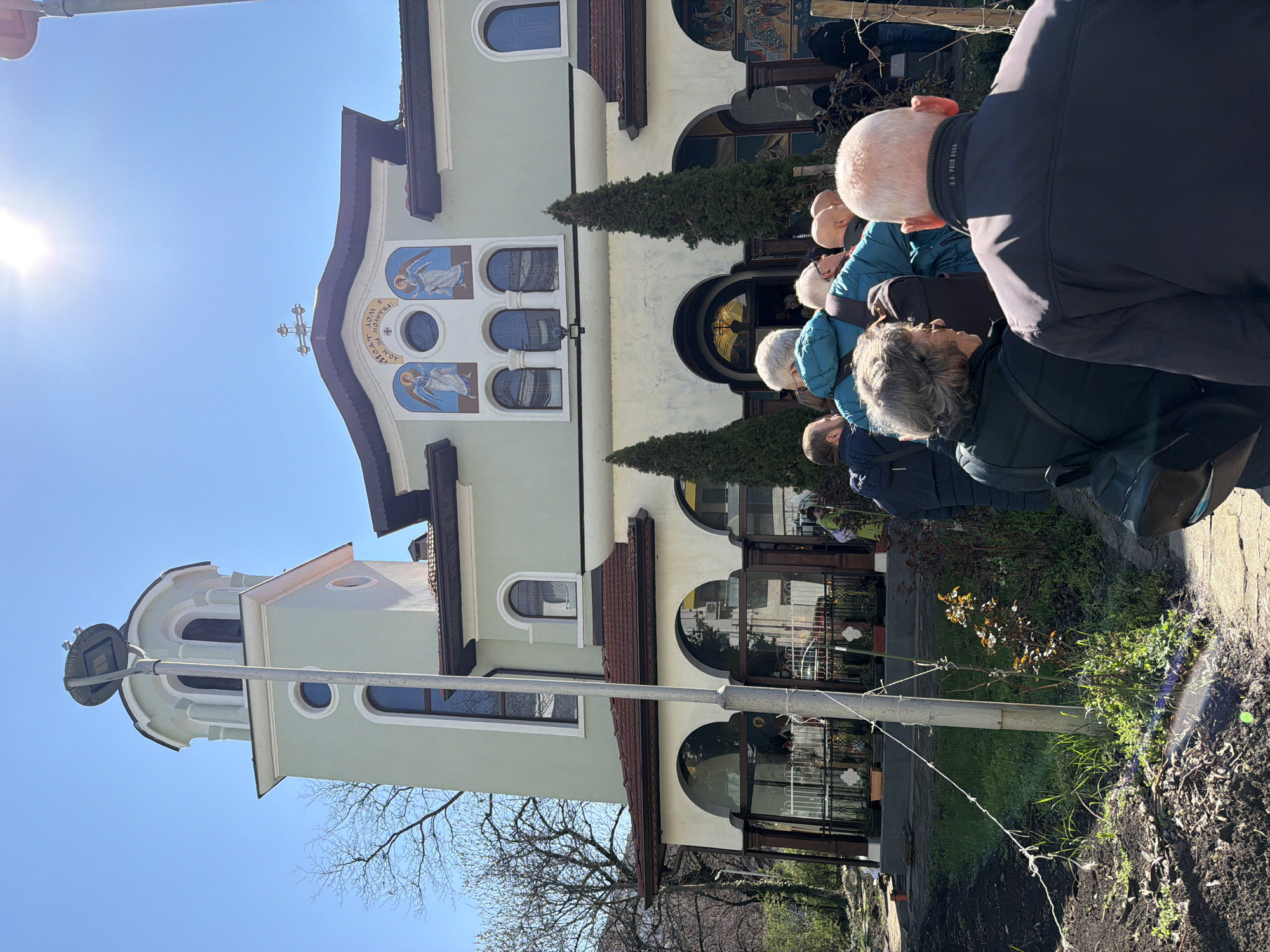 The image shows a church building with light green and white walls in Sofia, Bulgaria. A group of people are standing in front of the building. The church has a cross on the top. The building has arched windows and doors. The sky is blue, and the sun is shining.