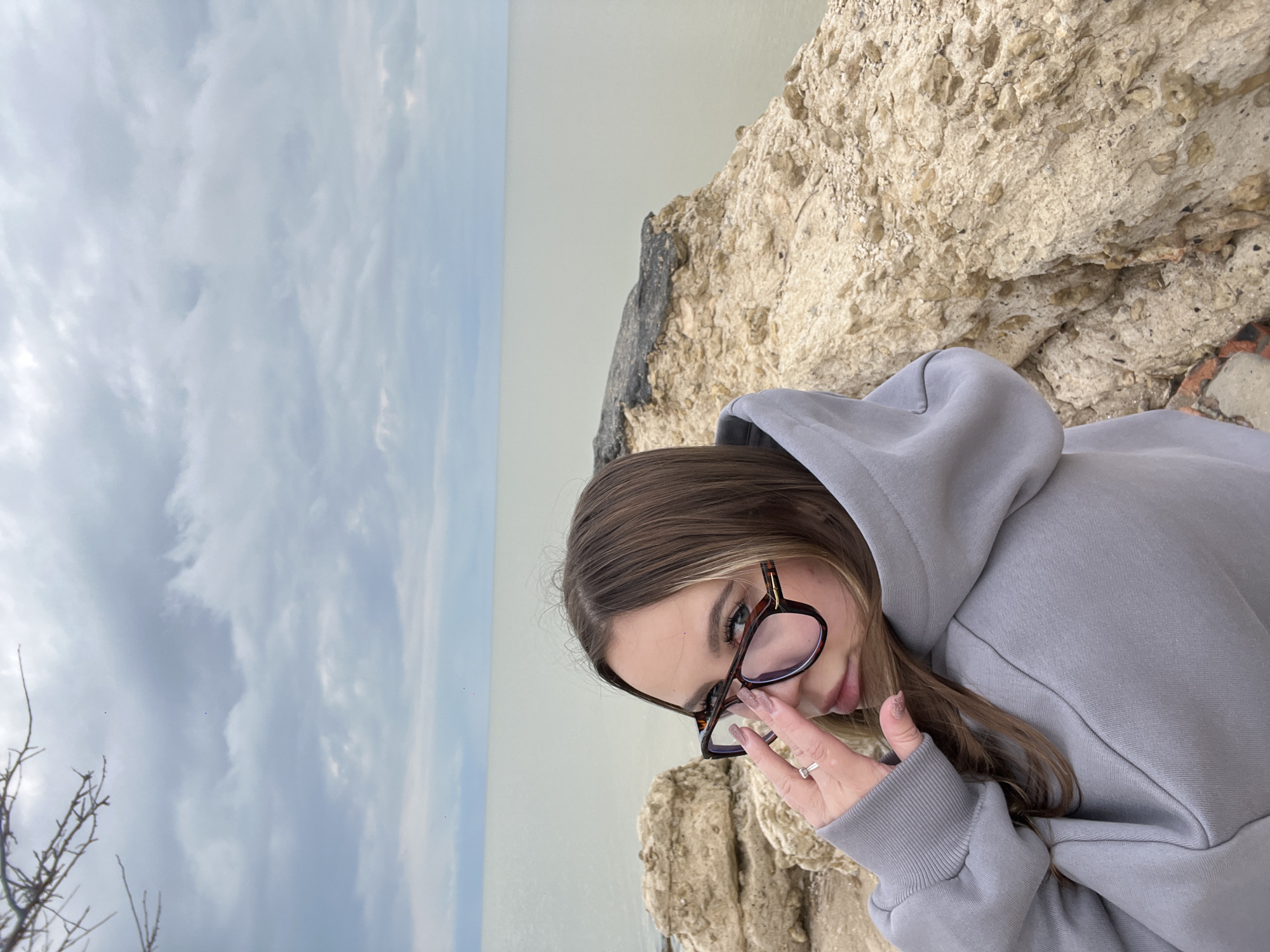 A young woman with long, light brown hair is pictured in an outdoor setting, her gaze directed towards the viewer. She is wearing a grey hooded sweatshirt and dark brown, possibly tortoise-shell, framed glasses, which she holds near her nose with her left hand. A silver ring is visible on her left ring finger. Her expression is neutral and direct.
The background features a textured, light-colored rock formation, indicative of a cliff or large boulder, occupying the right side of the frame. Beyond the rock and extending across the horizon is a calm body of water, likely the sea or a large lake, meeting an overcast sky filled with soft, diffused clouds. Some bare tree branches are visible in the lower left corner against the cloudy sky. The lighting suggests an overcast daytime. The location is contextualized as Port-Katon, Russia. There is no visible text in the image.