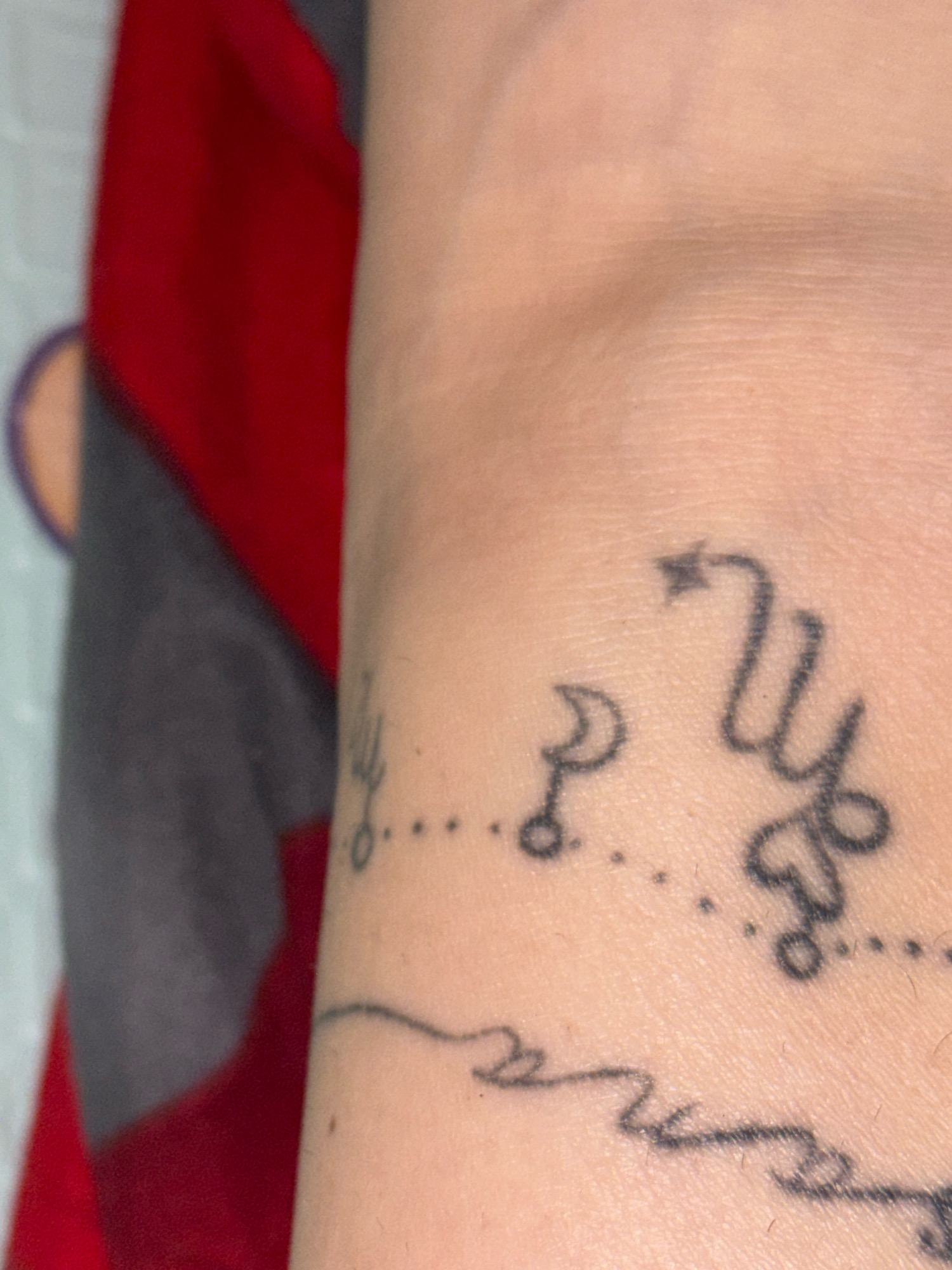 A close-up, horizontal shot displays a person's forearm with a tattoo of a Scorpio symbol and a crescent moon, connected by a dotted line. The skin is light-toned and shows some texture. A red and grey garment is visible in the background, suggesting the arm is resting against it. The overall lighting is soft, highlighting the details of the tattoo. There is no indication of the location being in Rusalevka, Russia, or any other specific city.