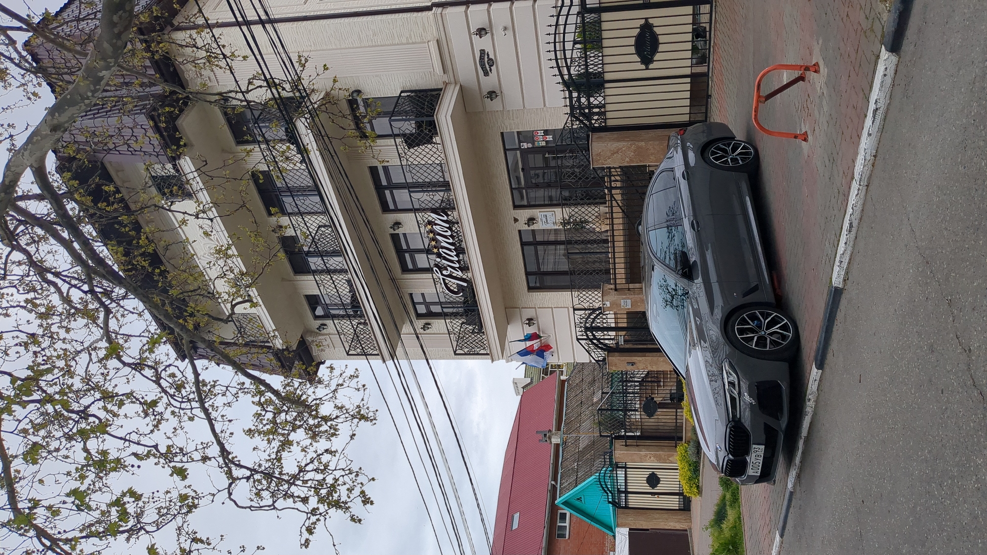 The image shows a five-story building identified by a sign as "Piano" (presumably a hotel), with a gray BMW car parked in front. The building is light-colored, with multiple windows, small wrought-iron balconies, and decorative railings. A tree with bare branches is present on the left side of the image. A red-roofed building is visible in the background. Power lines are strung across the sky. The license plate of the car appears to read "A005YB93," suggesting Russian registration. The scene is located in Gelendzhik, Russia.