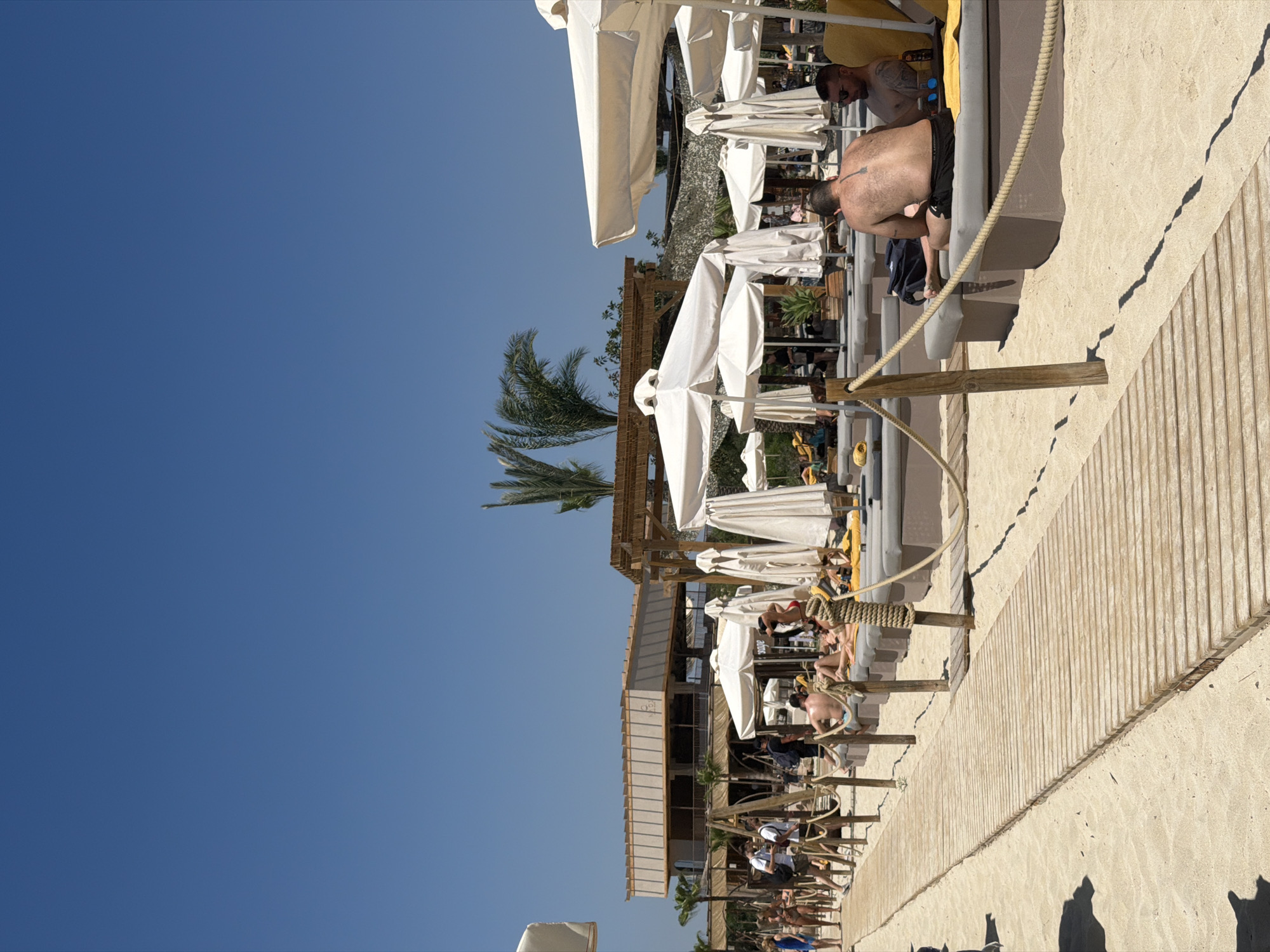 The image shows a sunny beach scene in Ayia Napa, Cyprus. A row of beach umbrellas and loungers are lined up along a wooden boardwalk. People are relaxing on the loungers. The sky is clear and blue. Palm trees can be seen among the umbrellas.