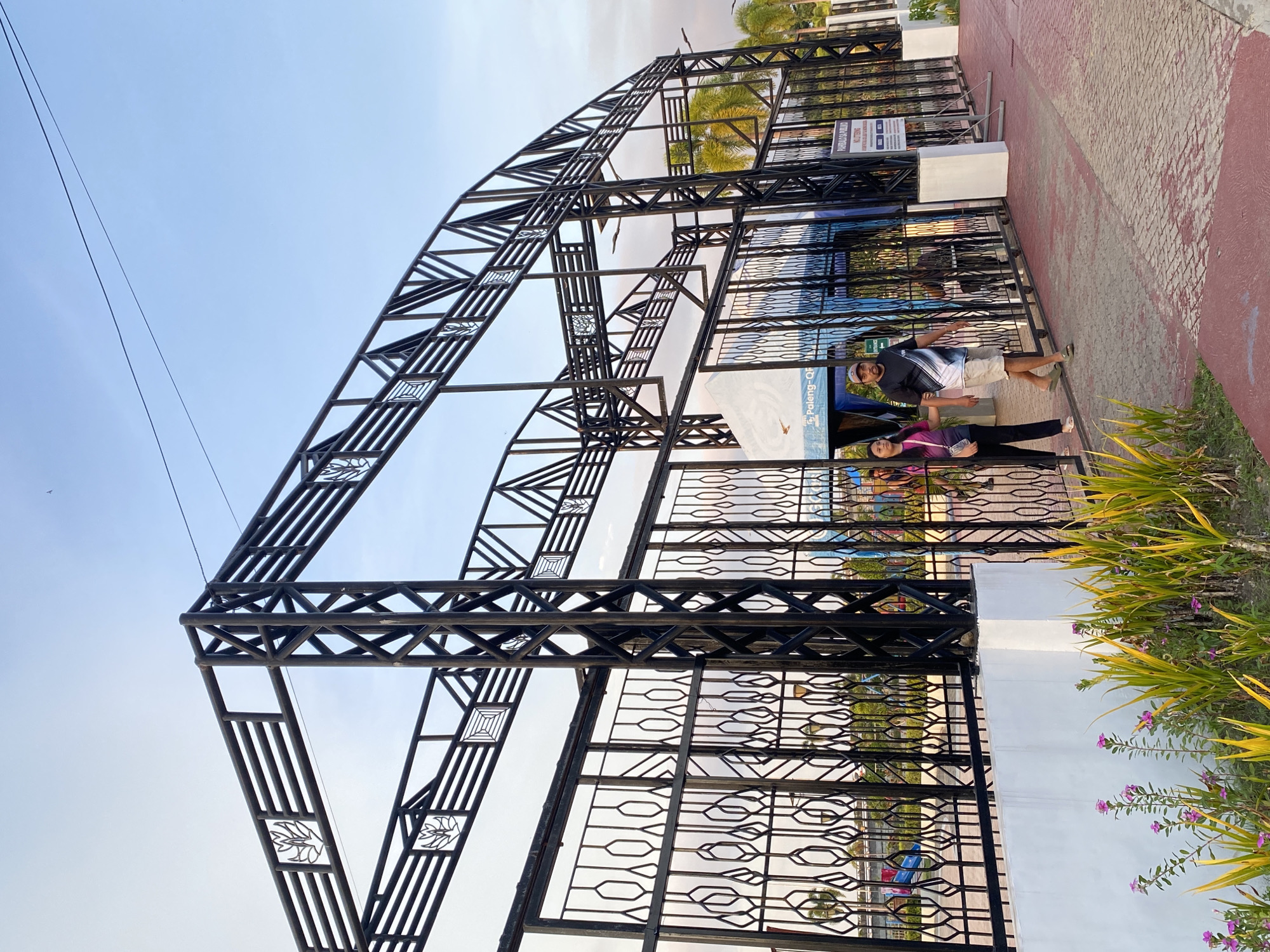This image captures an outdoor scene at the Davao City Hall Complex, Philippines. A large, black metal archway structure with intricate geometric patterns and decorative elements frames the entrance to a public space. The archway is adorned with stylized leaf motifs.
In the foreground, a paved walkway with a red and grey patterned surface leads towards the entrance. To the left, a patch of greenery with yellow and green foliage, interspersed with small pink flowers, lines the white base of a wall.
Two individuals are walking away from the viewer, towards the archway. A woman on the left, wearing a pink and black top and black pants, is walking with her arm extended backward. A man on the right, in a black and white patterned t-shirt and beige shorts, is walking next to her.
Behind the metal gates of the archway, a white tent structure is visible, bearing text that appears to read "Paleng-QF" and "ENTRANCE". Further in the background, palm trees suggest a tropical environment. A signpost with text in Filipino is also visible on the right side of the entrance. The sky above is a clear blue with a few faint clouds.