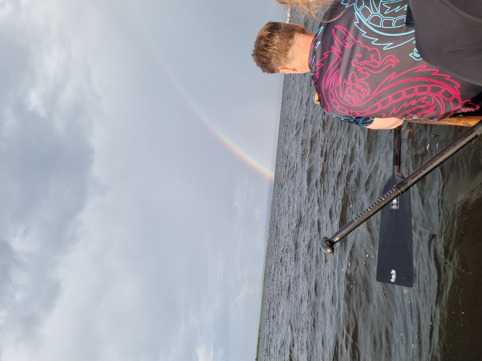 The image depicts a rear view of two individuals in a boat on the water, with a rainbow visible in the distance against a cloudy sky. The person in the foreground, facing away from the camera, is wearing a black t-shirt with a neon pink and blue dragon design. To their left, a paddle with a black blade is partially submerged in the dark, rippling water. The horizon is lined with a faint green strip of land, and in the far distance, some structures are visible. The overall atmosphere is overcast, suggesting recent rain or the approach of a storm. The location is identified as Chaikovsky, Russia.