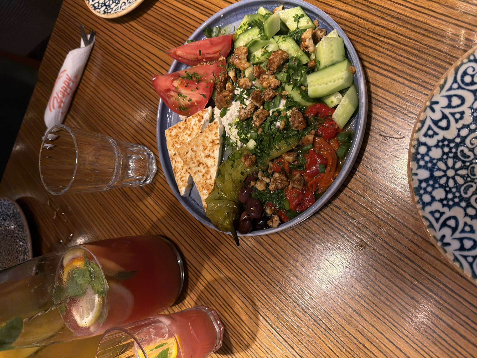 The image displays a meal set on a polished wooden table with distinct parallel grooves. A blue-grey ceramic plate in the center features a diverse assortment of food: two triangular pieces of pan-seared cheese are positioned at the top left, while two large red tomato wedges, garnished with chopped green herbs, are at the top right. A single green roasted pepper and a cluster of dark olives are situated on the mid-left of the plate. Below the cheese, a white creamy dip or spread is topped with chopped green herbs and scattered walnuts. To the left of the cucumbers, roasted red peppers are visible, also garnished with herbs and walnuts. Sliced cucumbers, similarly garnished, occupy the right side of the plate. To the left, a tall clear glass bottle holds a red beverage, garnished with fresh mint leaves and lemon slices, and is accompanied by a smaller glass containing the same drink. An empty clear glass tumbler stands above the main plate. In the upper right corner, a white rolled napkin bears the red script "Happy," with a metal fork partially visible inside. Sections of two ceramic plates, patterned in blue and white, are visible in the top left and bottom right corners of the scene. The lighting produces subtle reflections on the table surface. This scene is identified as taking place within a Happy Bar & Grill restaurant, a chain with multiple establishments in Sofia, Bulgaria, evidenced by the "Happy" branding on the napkin.