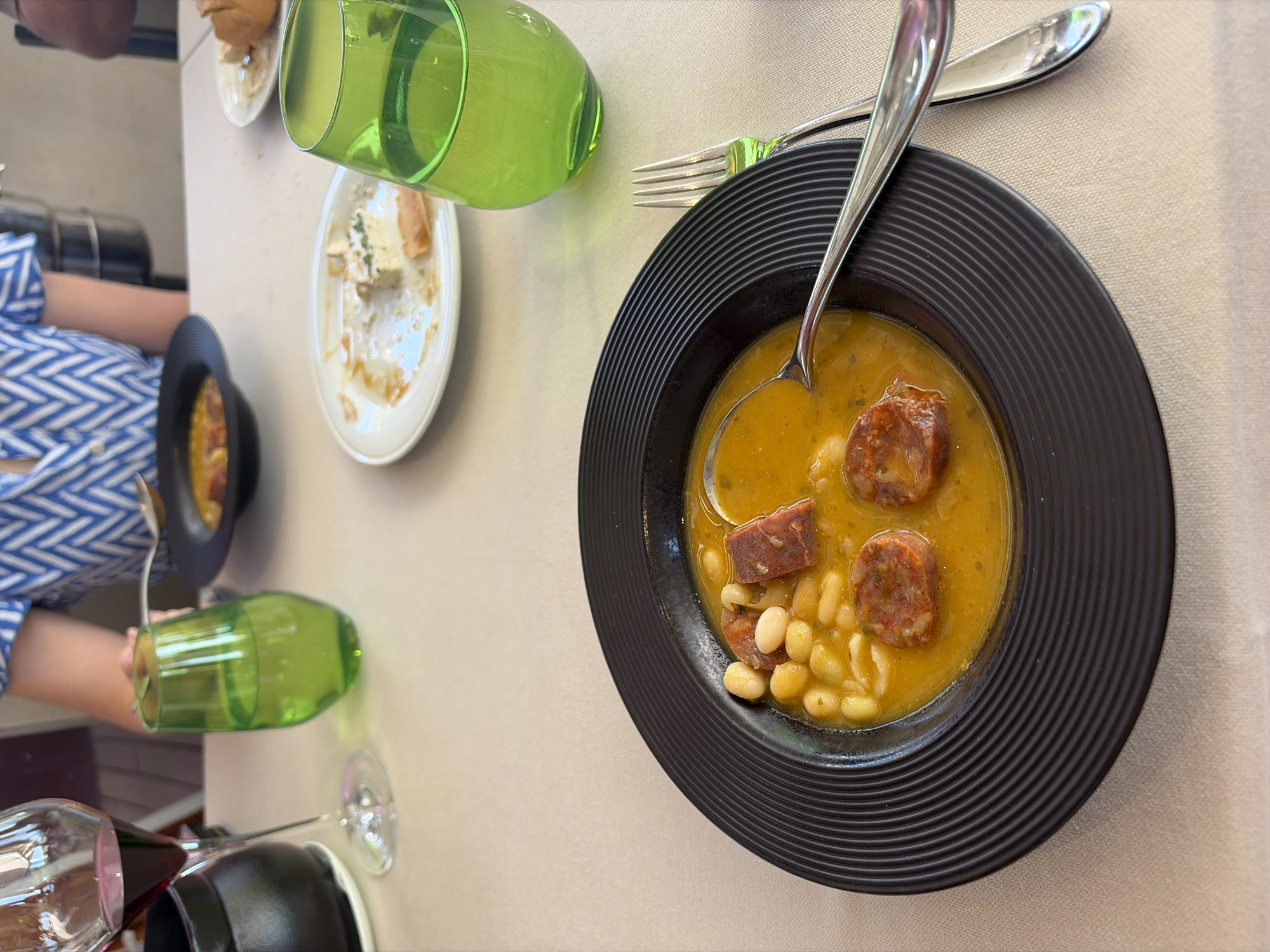 The scene depicts a table setting in Elciego, Spain. A person wearing a blue and white patterned shirt is seated at the table. On the table, there's a black bowl filled with a stew-like dish, containing beans and what appears to be sausage, with a spoon placed inside. A green glass is overturned near the bowl. To the side, there are plates, one clean and one partially eaten, along with some bread. Silverware (fork and knife) are neatly arranged. The overall environment suggests a dining experience. A wine glass is visible near the bottom of the image.