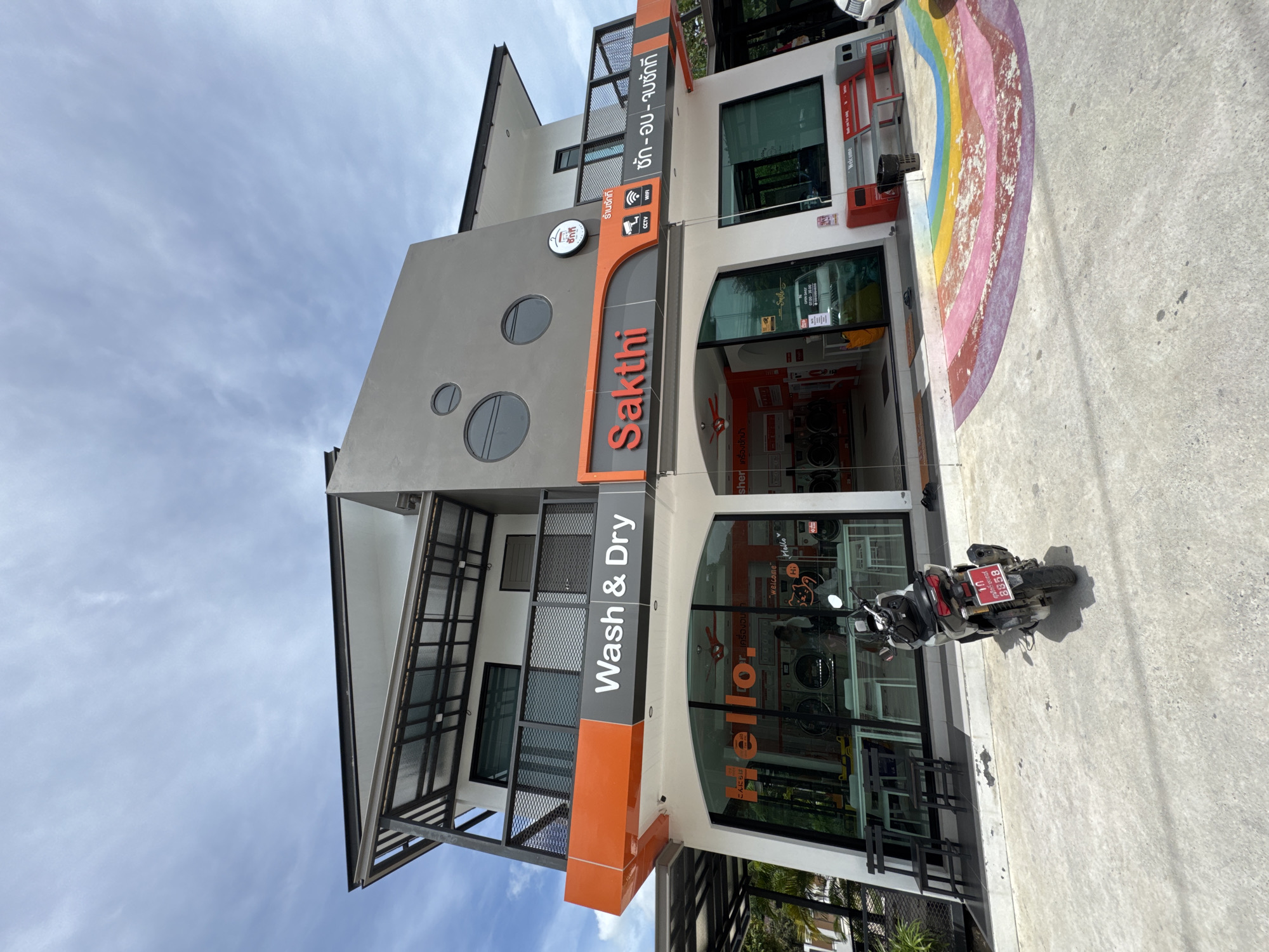 A sideways shot depicts the exterior of "Sakthi Wash & Dry", a laundromat located in Moo 4, Thailand. The modern, two-story building features a gray facade with circular windows and an orange vertical sign with the business name written in English and Thai, along with "Wash & Dry". Large glass windows reveal orange washing machines inside. A motorcycle with a Thai license plate is parked in front. A faded rainbow design is painted on the ground adjacent to the building. The sky above is blue with scattered clouds.