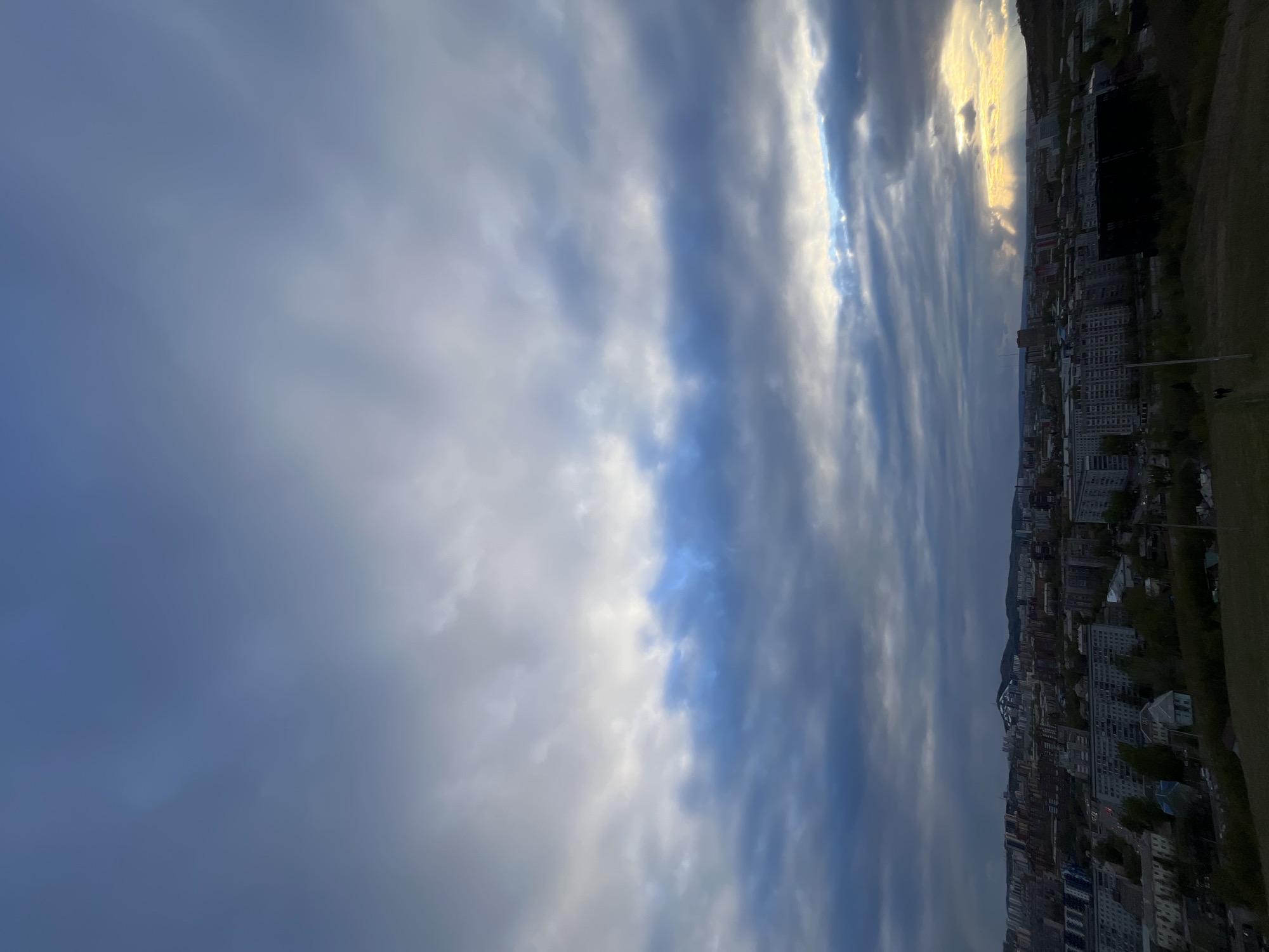 The image shows a cityscape in Krasnoyarsk, Russia, under a dramatic sky. The city consists of high-rise buildings of varying heights and colors, suggesting residential and commercial areas. The buildings stretch towards the horizon, where the landscape rises into a hill or mountain. The sky is dominated by layered clouds in shades of white, gray, and blue, with a patch of sunlight breaking through near the top right of the frame. A grassy verge occupies the top right corner, indicating the vantage point is elevated. No people are clearly visible, but the urban landscape suggests human activity.