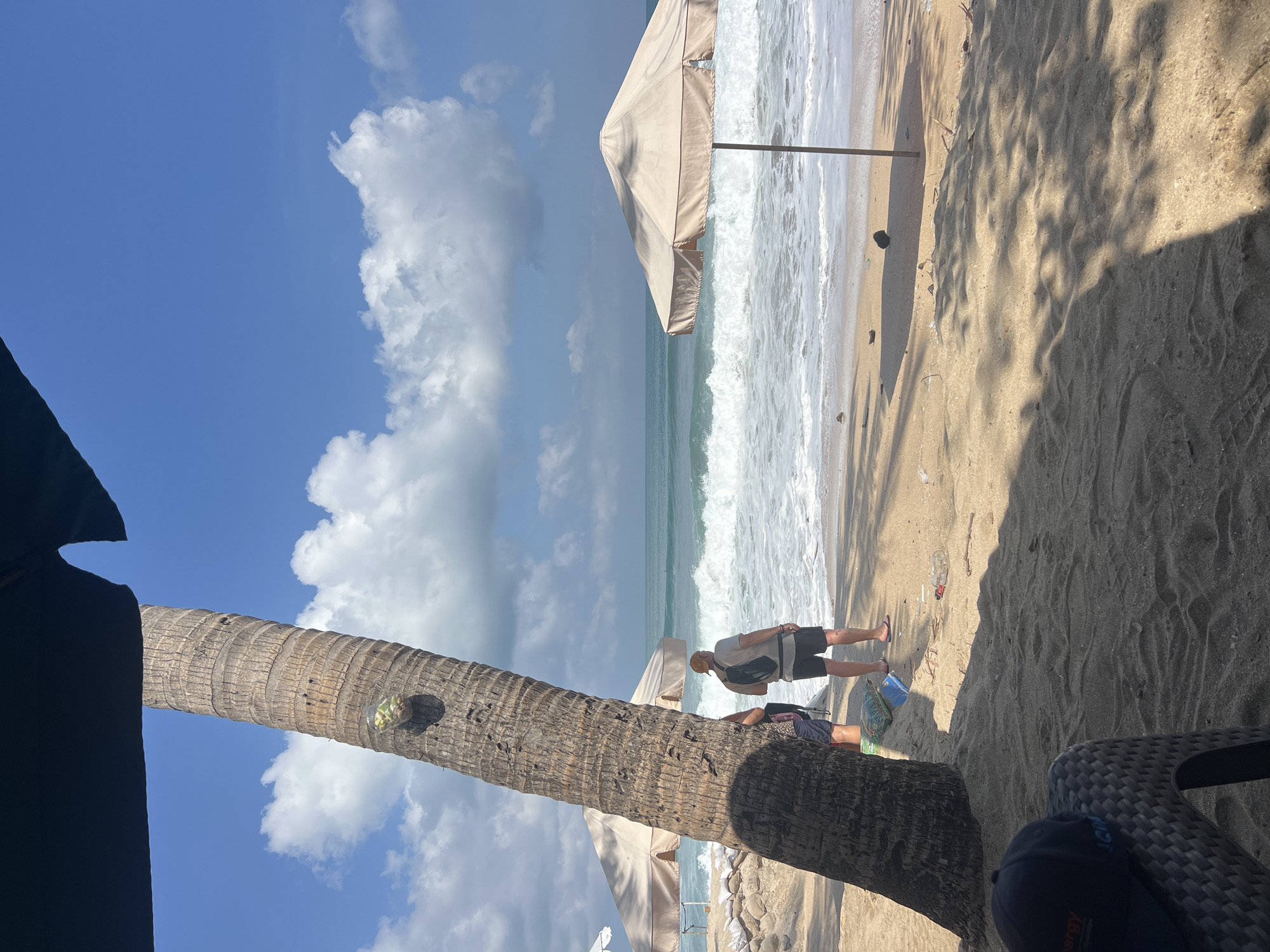 This image depicts a sunny beach scene in Kuta, Indonesia. In the foreground, a large palm tree trunk is prominently featured on the left, partially obscuring the view. A black umbrella casts a shadow over the sandy ground.
Two individuals are standing on the beach near the waterline. One person, wearing a grey t-shirt, black shorts, and a cap, faces the ocean, carrying a backpack. Another person, partially visible, is standing next to them.
Further back, a beige beach umbrella is set up. The ocean displays moderate waves with white foam crashing onto the shore. The sky is a clear blue, dotted with fluffy white clouds. Scattered on the sand are some plastic bottles and debris, along with coconuts. A blue bag and a brown item are also visible on the sand. In the far left, another beach umbrella is visible. The foreground shows a dark blue cap on a woven chair.