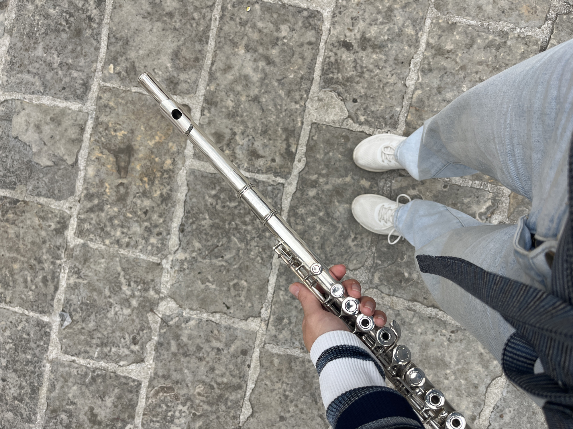 A person wearing light blue jeans and white sneakers stands on a paved stone surface, holding a silver flute in their hand. The person's arm is visible, clad in a white and navy striped sweater. The background is a paved surface made of grey stone blocks. The location appears to be an urban outdoor setting. No identifying landmarks are visible to confirm the specific location of Budva, Montenegro.