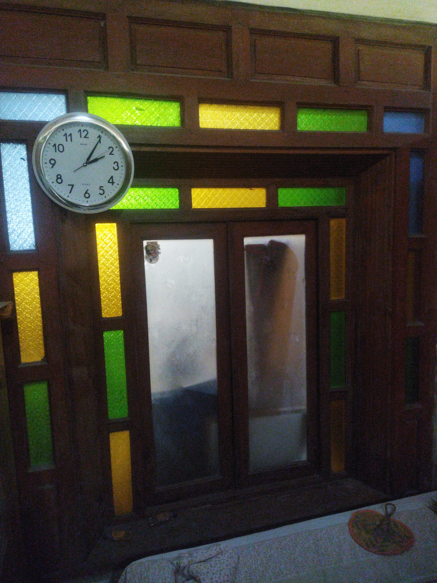 This is a medium shot, eye-level photograph of a traditional Javanese window and clock. The window is made of dark wood and features multiple panes of colored glass in shades of green, yellow, and blue. The glass is translucent, obscuring the view through the window. A round analog clock with a white face and black numbers is mounted to the left of the window. The clock hands indicate the time is approximately 1:55. The overall scene appears to be indoors, with a textured, possibly fabric surface visible in the lower portion of the frame. The lighting is dim, with reflections visible on the glass panes. The image is grainy, suggesting it was taken in low light conditions or with a lower quality camera. The location is specified as Juwiring, Indonesia, implying a cultural context for the architectural style and decorative elements.