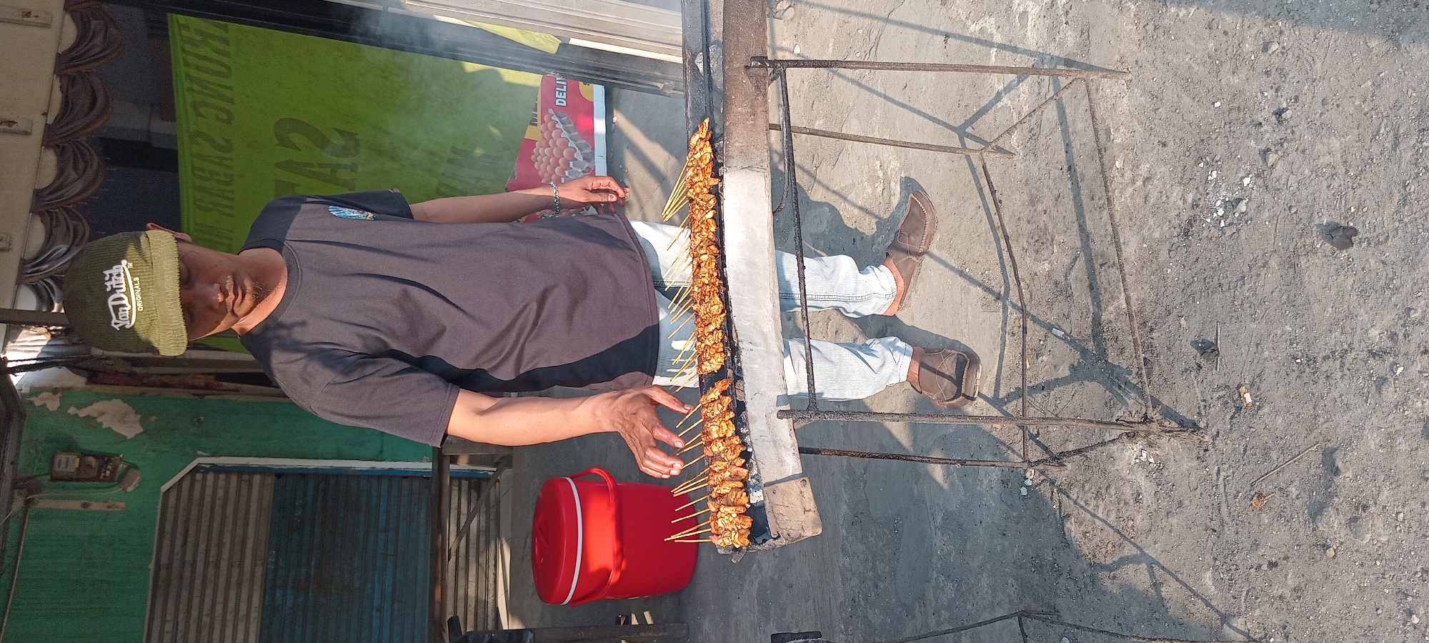 A man wearing a dark t-shirt, light blue jeans, brown loafers, and a green baseball cap with "Van Dutch ORIGINALS" embroidered on it, is standing in front of a grill laden with rows of uncooked satay skewers. He is positioned to the left of the grill, with his right hand reaching towards the skewers. To the left of the man and grill, a bright red cooler is visible. The scene appears to be outdoors, with a concrete ground surface. In the background, a green wall and a shuttered door are present, along with a faded yellow banner displaying text in Indonesian that reads "TRUNG SABAR IN SAZ DELI". The image captures a moment of food preparation at what appears to be a food stall or roadside eatery in the city of Karangsari, Indonesia.