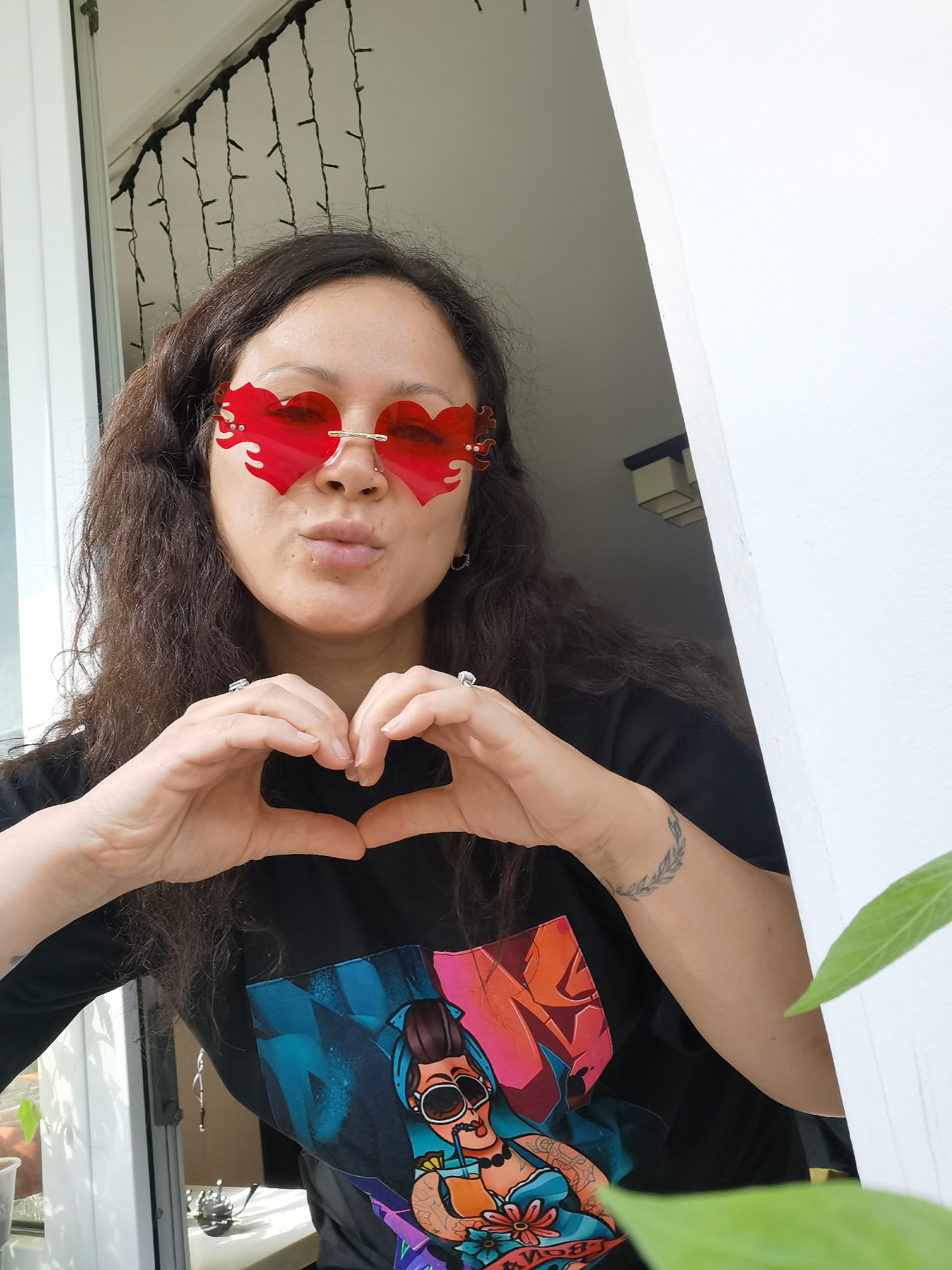A woman with dark, curly hair and red heart-shaped glasses stands in a white-framed doorway, making a heart shape with her hands. She wears a black t-shirt with a colorful graphic print. A tattoo is visible on her left wrist. Behind her, strings of lights hang in front of a window. To the right, a green leaf is partially visible. The location appears to be indoors, looking out towards natural light. The city of Chaikovsky, Russia is not explicitly identifiable in the image.