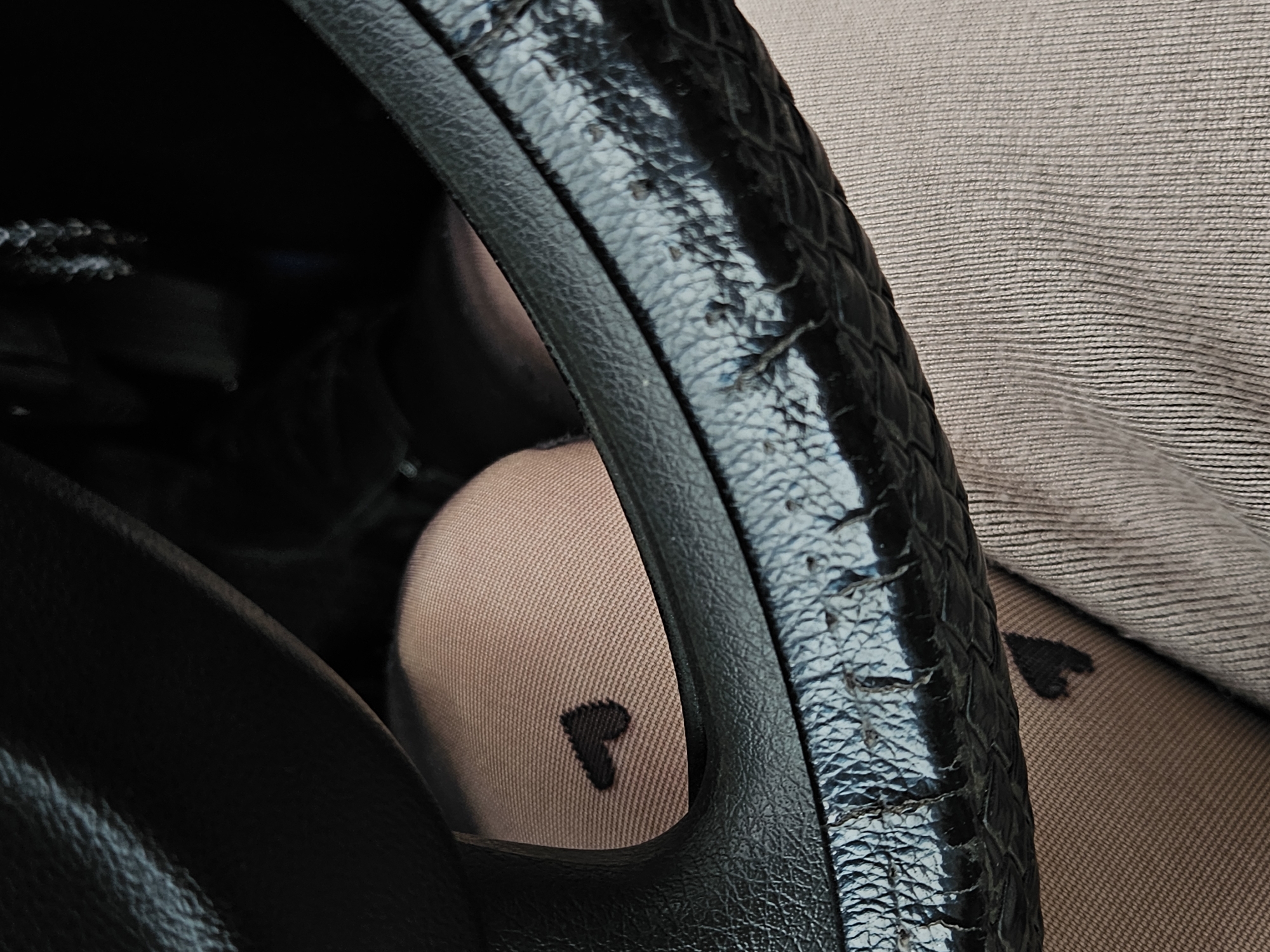 The image shows a close-up of a worn, black, reptile-textured steering wheel in an automobile. A person's leg, clad in light-colored tights with a small, dark heart pattern, is visible behind the steering wheel. Part of the car's dashboard or interior is also visible in the background. There is no location in Chaikovsky, Russia, identified.