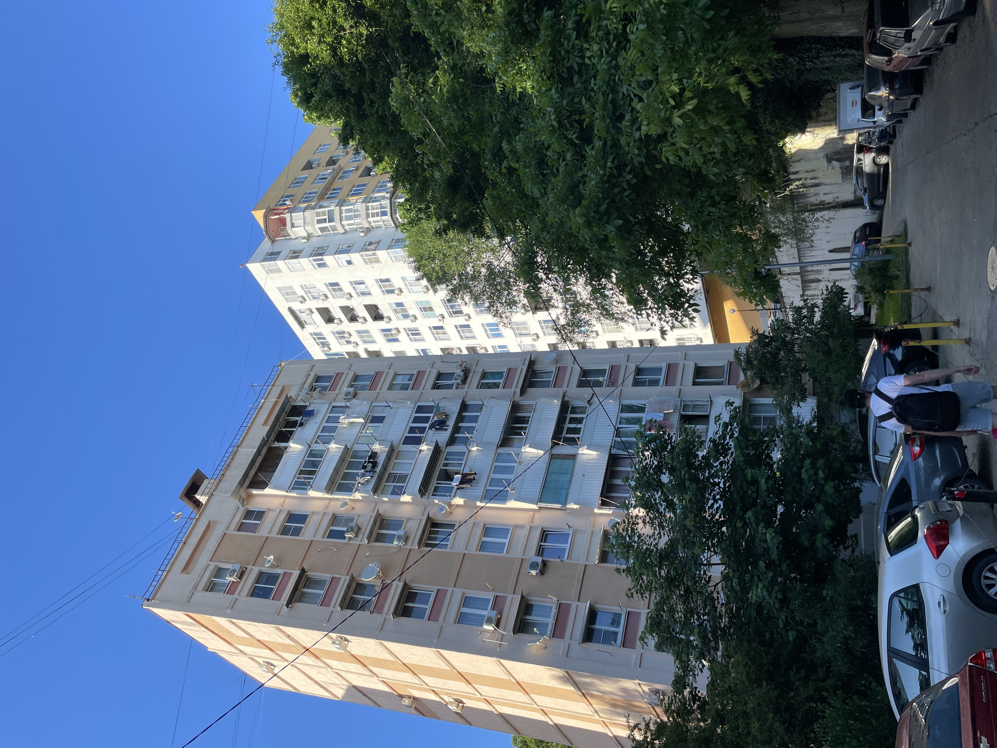 This image depicts an urban residential scene in Sochi, Russia, captured under a clear blue sky. The foreground features a paved pathway or narrow street with numerous parked cars of various models and colors lining both sides, including a silver hatchback, a dark sedan, and several older vehicles. Yellow bollards delineate parts of the pedestrian area.
To the left, a tall, multi-story residential building with a beige and light brown facade dominates the scene. Its exterior is marked by numerous white-framed windows, individual air conditioning units, and several satellite dishes. Clothes are visibly drying on some of its balconies. Power lines traverse the upper portion of the frame, near the building's roof which features a small structure and a railing.
To the right of the beige building, and partially obscured by dense green foliage, stands another tall residential building. This structure is predominantly white and light yellow, featuring numerous windows and distinct curved balconies on its corner section. The lush green canopy of several mature trees occupies a significant portion of the midground, partially concealing the buildings and casting shadows on the ground.
One individual, seen from behind, walks away from the camera on the paved path. This person is wearing a white t-shirt, light-colored shorts, and a black backpack. Further down the street, beyond the parked cars, a retaining wall or building foundation is visible, overgrown with some greenery in parts, and a white commercial vehicle is parked. The strong sunlight and distinct shadows suggest a daytime setting, likely late morning or early afternoon.