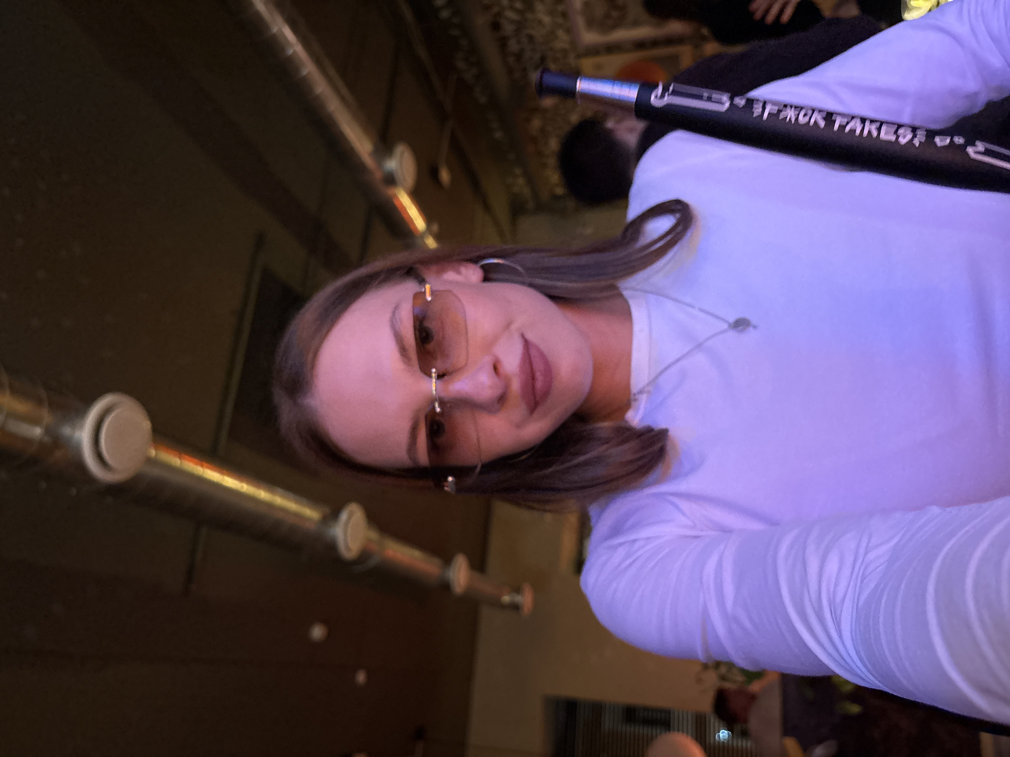 A young woman with shoulder-length brown hair and rimless rectangular sunglasses is captured in a selfie, illuminated by purple lighting. She is wearing a long-sleeved white shirt and a delicate silver necklace. She is holding a black object with white text that appears to be a hookah hose, draped over her shoulder. The background suggests an indoor venue with exposed ductwork on the ceiling and a dimly lit atmosphere. People are faintly visible in the background, blurred by the low light. The text on the hookah hose is in Cyrillic and reads "КАЛЬЯН TAKEOFF" which translates to "Hookah TAKEOFF". This suggests the location is likely a hookah lounge or a bar in Perm, Russia, potentially named "Hookah TAKEOFF".