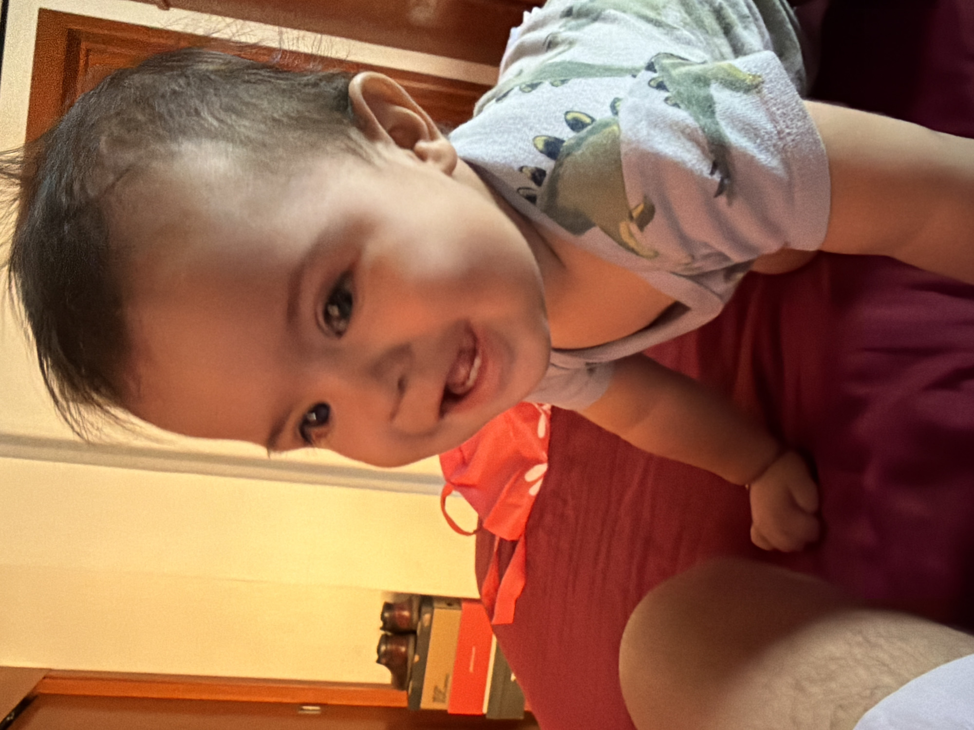 The image shows a baby lying on a burgundy surface. The baby is wearing a light-colored onesie with dinosaur patterns. The baby is looking towards the camera with a slight smile, revealing their teeth. A portion of an adult's leg is visible in the lower right corner of the image. In the background, a wooden door, a shelf with books and boxes, and a wall with yellow lighting are present. The image was not taken in Reforma, Mexico. There is nothing in the image to indicate that it was taken in Reforma, Mexico.