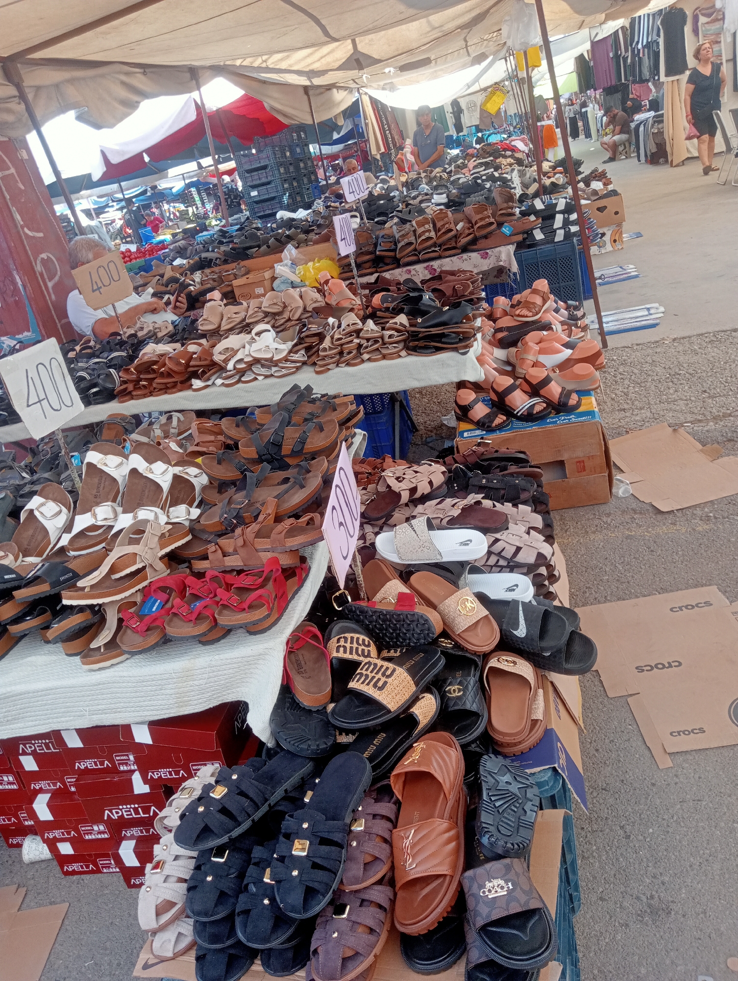 This is a daytime, eye-level, outdoor photograph of a bustling street market in Antalya, Turkey, likely a bazaar. The foreground is dominated by a large display of footwear, primarily sandals and flip-flops, neatly arranged on tables and boxes. The sandals are diverse in style, color, and material, with many appearing to be leather or faux leather. Some pairs are displayed with price tags, one prominently showing "400" and another "300".
In the background, more stalls are visible, selling clothing and other goods under white and red awnings. Several people are present: a man with grey hair is sitting behind a table of sandals, appearing to be a vendor, and other shoppers are browsing the stalls. The ground is paved with a grey, textured surface, and cardboard boxes are scattered around the area. The overall atmosphere suggests a vibrant commercial environment typical of a Turkish market.
