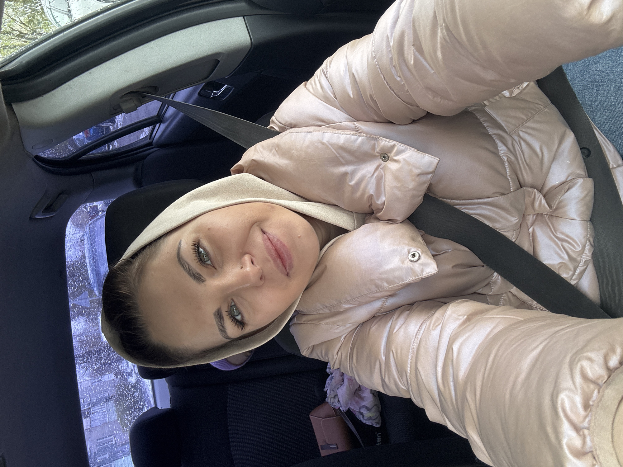 A young woman is shown in a selfie taken inside a car, likely in Раздолье, Russia. She is positioned in what appears to be the front passenger seat, wearing a light-colored, possibly rose gold or champagne, shiny puffer jacket over a beige hooded sweatshirt. A dark seatbelt crosses her chest diagonally. She has light green or hazel eyes and a gentle, pleasant smile, looking directly at the camera. Her dark hair is pulled back, and her face shows slight freckles or moles.
The car's interior is predominantly dark, with black fabric seats. Through the car's sunroof and side windows, an outdoor scene is visible, appearing blurred and abstract, possibly due to rain or snow on the glass, or an artistic effect. The blurred elements suggest trees and possibly buildings in the background. The lighting indicates it is daytime, perhaps an overcast day, given the soft, diffused light.
Next to her on the car seat, a small, light brown or pink purse is visible, along with a patterned scarf or piece of fabric in pastel colors. There is faint, illegible text on the purse and a small white tag near it. The overall impression is a casual, in-car portrait.