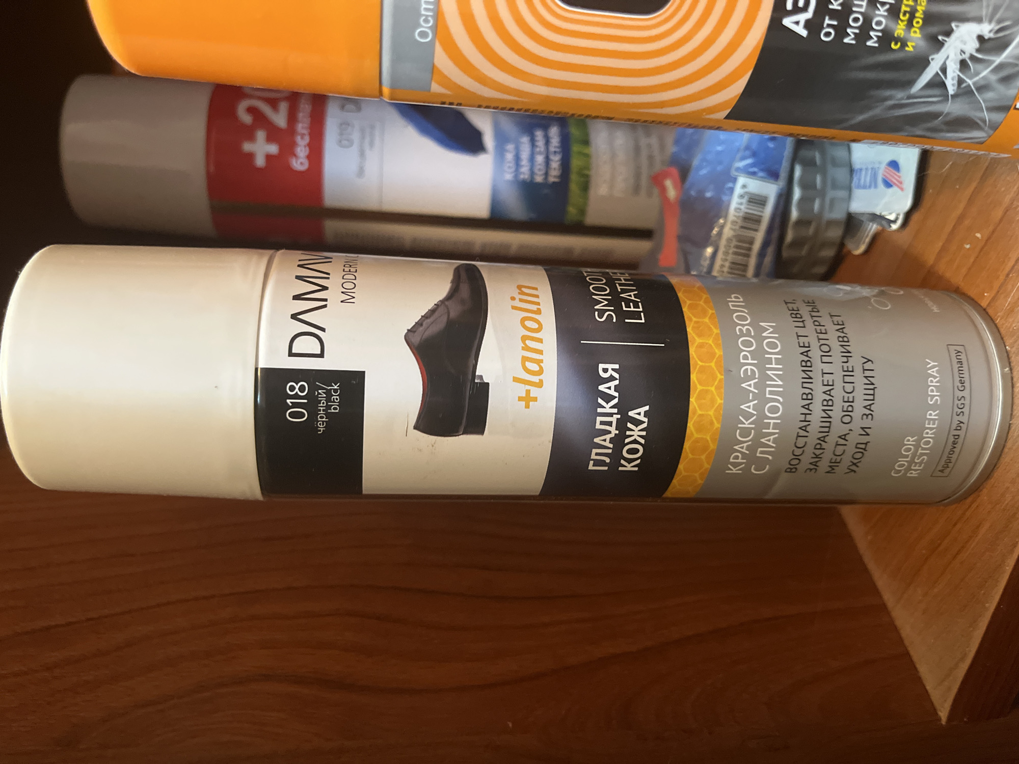 The image shows a close-up view of three aerosol cans on a wooden shelf. The primary focus is on the leftmost can, which is white and black, labeled "018", " DAMAV" and "MODERN". It features an illustration of a black dress shoe and the text "+lanolin" and "ГЛАДКАЯ КОЖА SMOOTH LEATHER". Further down, it says "КРАСКА-АЭРОЗОЛЬ С ЛАНОЛИНОМ". The middle can is mostly out of focus, with a red and white label. The rightmost can is bright orange and white with a patterned design and text that is partially obscured but appears to be an insect repellent. The background is a dark wooden surface, possibly a cabinet or shelf. The lighting suggests an indoor environment. There are no people or animals visible in the image. The scene is static. There is no information in the image to suggest its location in Minsk, Belarus.