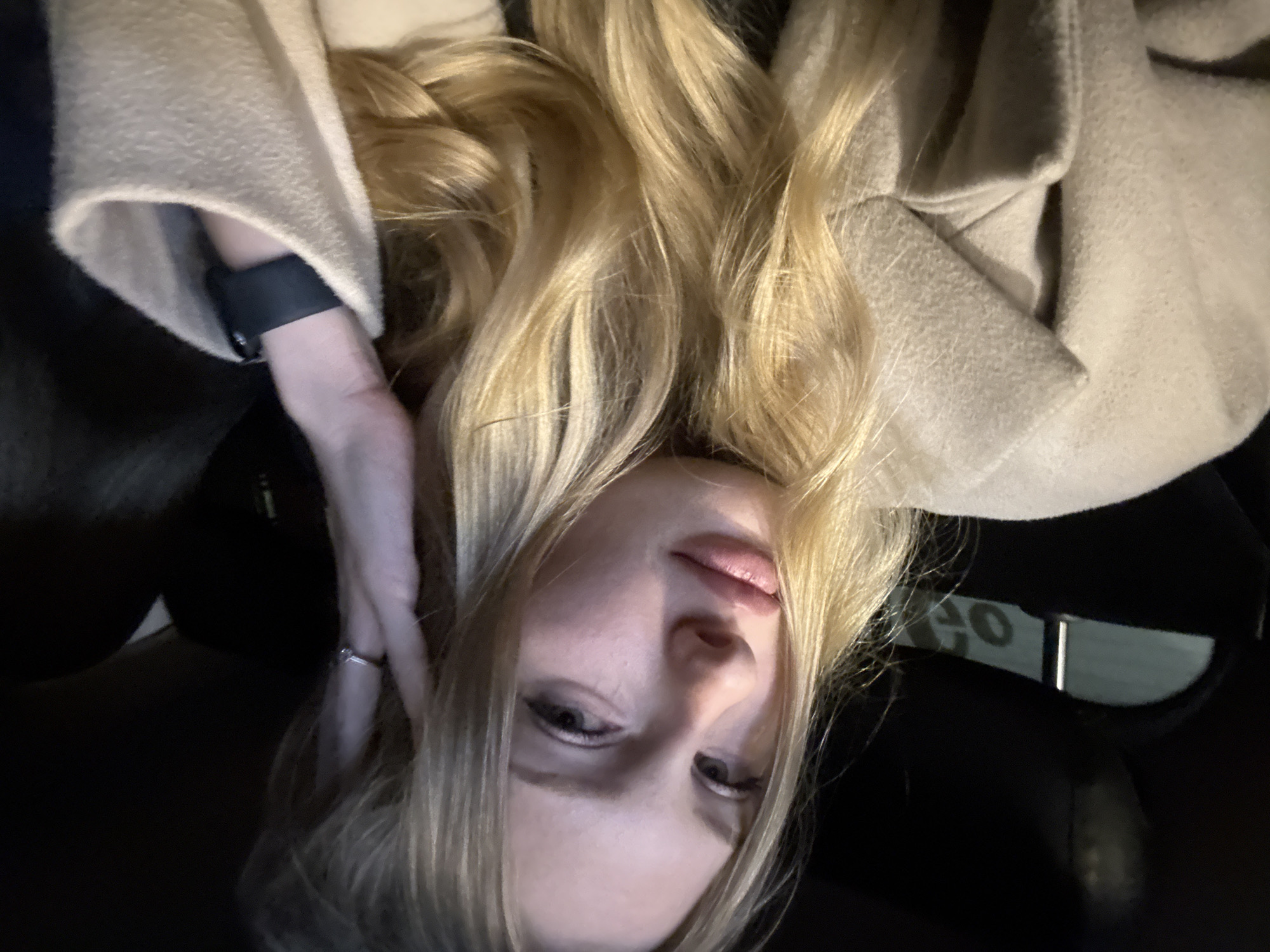 A young Caucasian woman with long blonde hair is seated in the interior of a vehicle. She is wearing a beige coat and a dark smartwatch on her left wrist. Her right hand is raised to her head, and she is looking towards the camera with a relaxed expression. The background is dark, suggesting the interior of a car at night or in low light conditions. A blurred object with the letters "GO" is visible in the background on the left side of the image, which could be signage from a business or street. There is no indication in the image that the location is specifically the city of Perm, Russia.