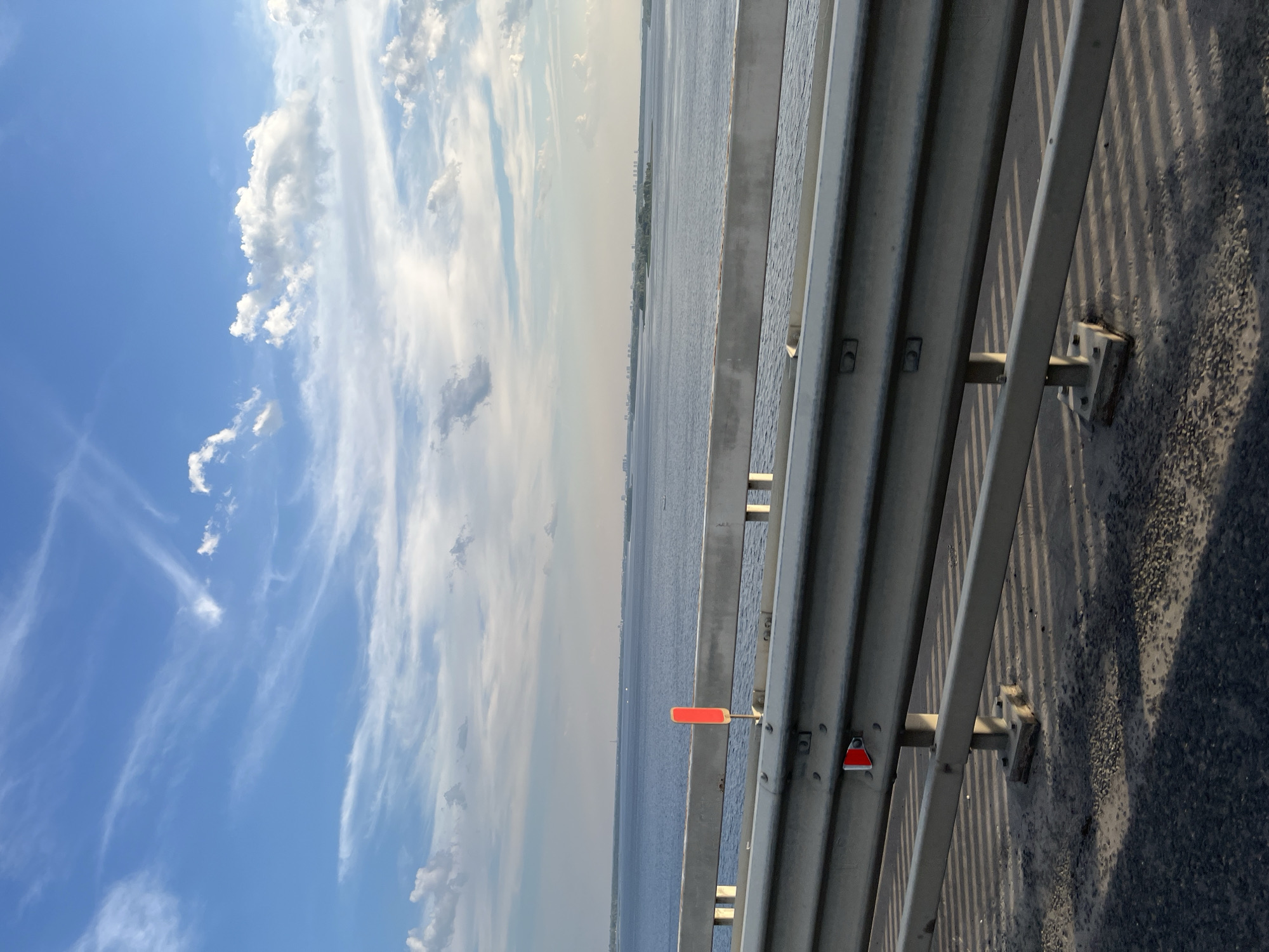 The image is a view from a bridge overlooking a wide, calm body of water under a bright blue sky with scattered white clouds. A silver metal guardrail runs horizontally across the foreground of the image, casting long, striped shadows onto the asphalt of the bridge. There are two red reflectors visible on the guardrail. In the distance, a long, low shoreline with buildings is visible, suggesting an urban or semi-urban environment. The water extends to the horizon, and the lighting suggests it is late afternoon or early evening. This scene is likely on the Volga River near the village of Pristannoye, Russia, given the expansive river and distant cityscape.