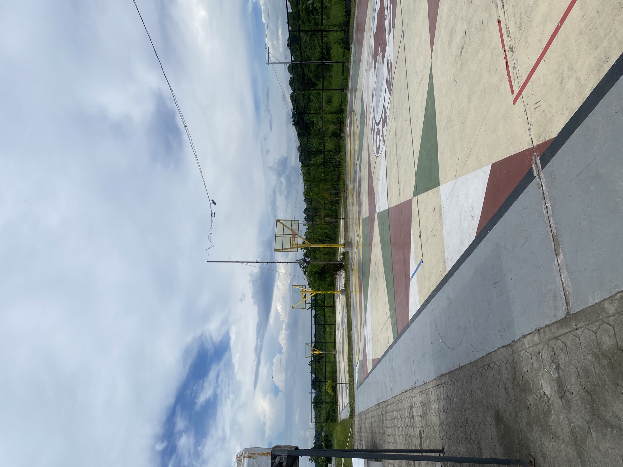 The image depicts an outdoor basketball court with colorful geometric patterns painted on its surface, located in Davao City, Philippines. Two basketball hoops with yellow poles and clear backboards are visible on the court. In the background, a black fence separates the court from a lush green area with trees and vegetation. The sky is partly cloudy with scattered white clouds against a pale blue backdrop. A few power lines are strung across the upper portion of the image. To the left of the court, a gray concrete walkway with hexagonal paving stones is present. The court appears to be part of a public or recreational facility.