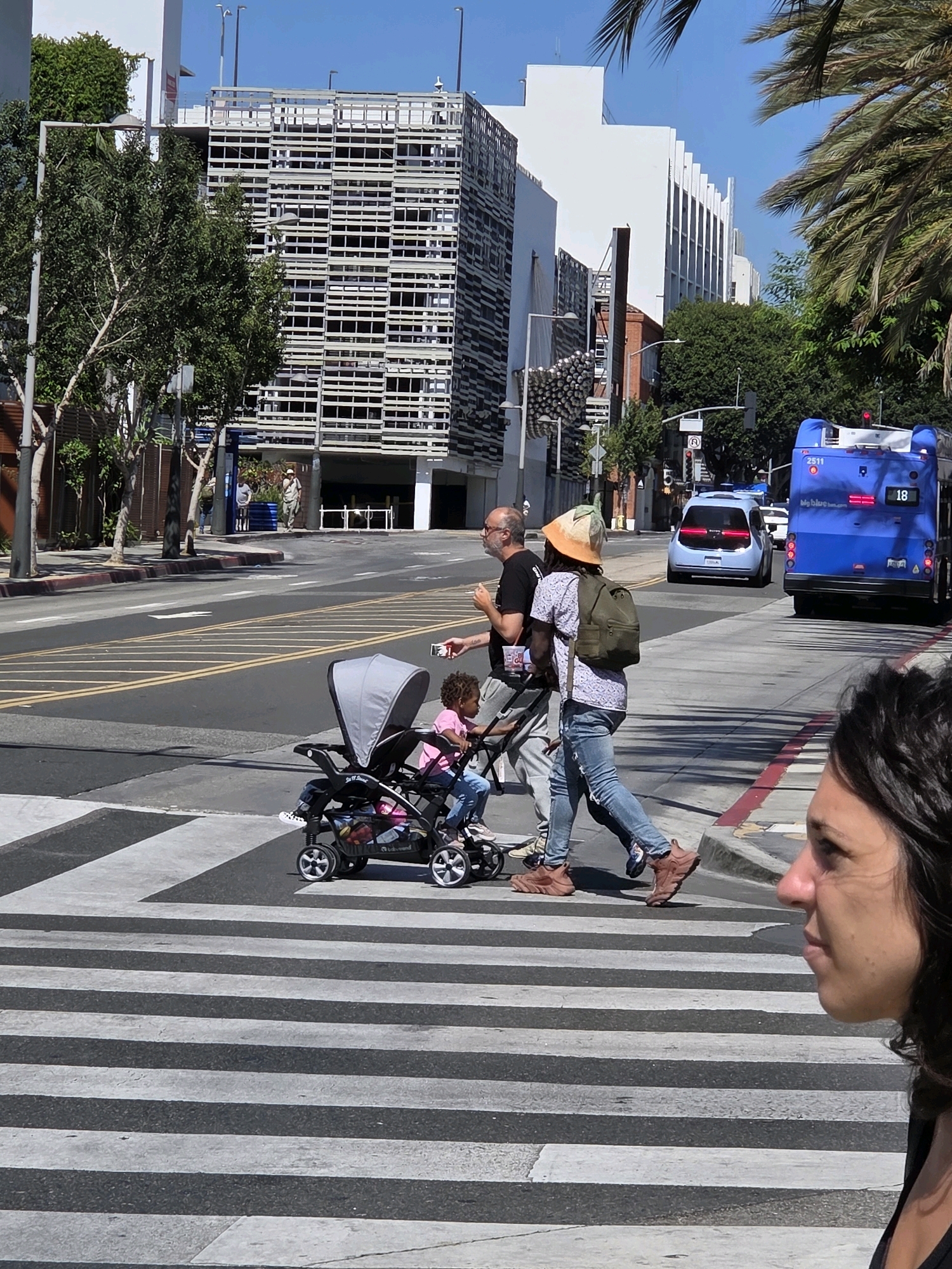 In Santa Monica, USA, a group of pedestrians crosses a street on a sunny day. In the foreground, the head and shoulders of a woman are visible on the right side of the frame, facing left. She has dark hair and is looking towards the street. Further back, a man wearing a black t-shirt and glasses walks with a stroller containing a young child. The stroller is gray with black accents and the child is wearing a pink shirt and blue jeans. Behind them, another person with a distinctive hat and backpack crosses the street. A bright blue bus is visible in the background on the right, with the number "18" displayed on its side. Several cars are also present on the road. The urban environment features modern buildings and palm trees, suggesting a warm climate. The pedestrians are walking on a marked crosswalk with black and white stripes.