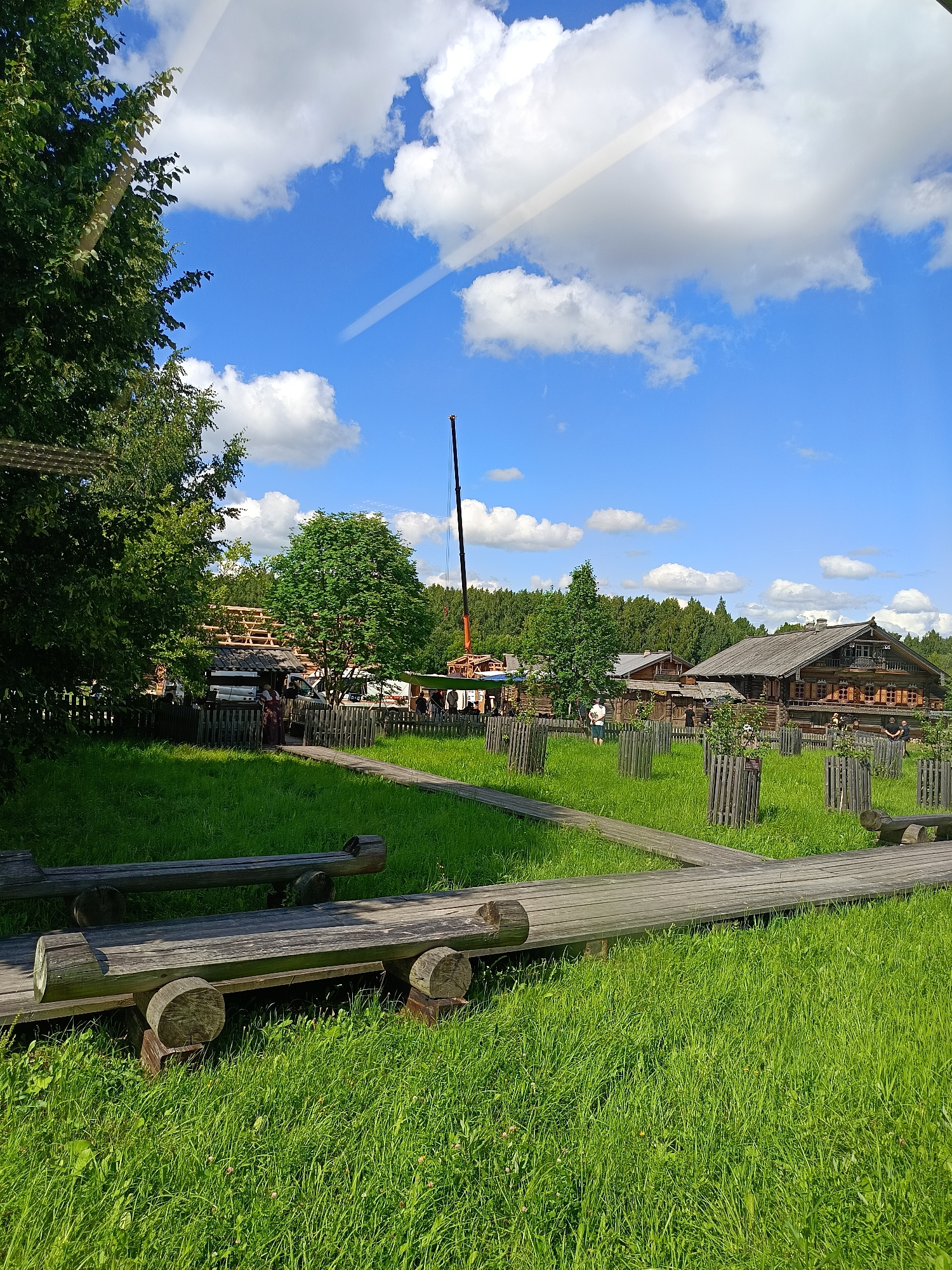 This is a medium-shot, outdoor, eye-level, vertical photograph taken on a sunny day in the city of Semenkovo, Russia. The scene depicts a village with several wooden buildings and a construction site in the background, a crane is visible. In the foreground, there are wooden benches and a wooden walkway path that leads through a grassy area with small trees enclosed in wooden fences. People are scattered throughout the scene, some are observing the construction, others are walking. The sky is blue with scattered white clouds.