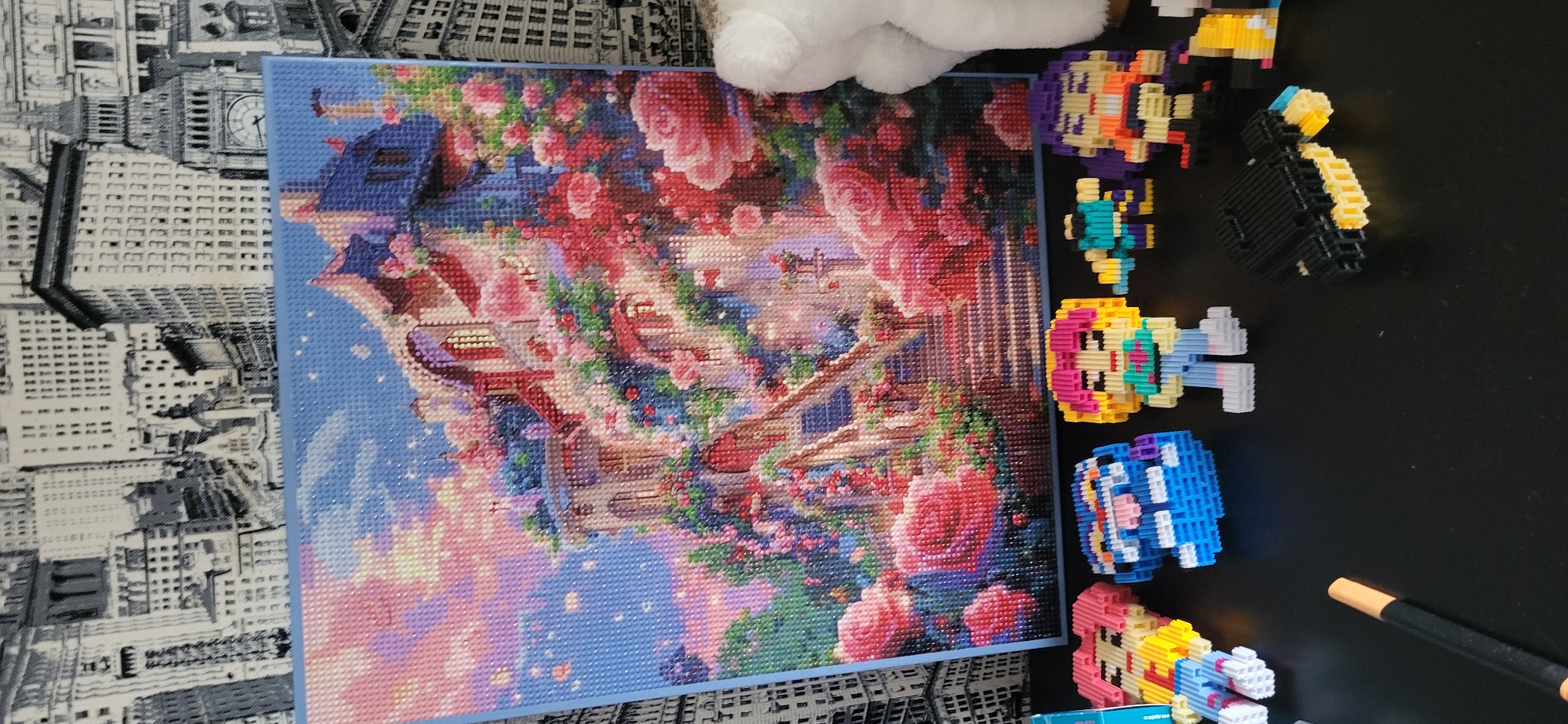 The image shows a close-up view of a diamond painting featuring a colorful, fantastical building adorned with roses against a starry, pastel sky. In front of the painting, several small, pixelated figures are arranged on a dark, reflective surface. These figures appear to be small, brick-built characters with bright colors and simple designs, resembling a collection of toys. To the left of the figures, a black marker or pen lies on the surface. The background of the image is a patterned wallpaper depicting a city skyline with tall buildings. There is no identifiable information within the image that places it in Oskemen, Kazakhstan.