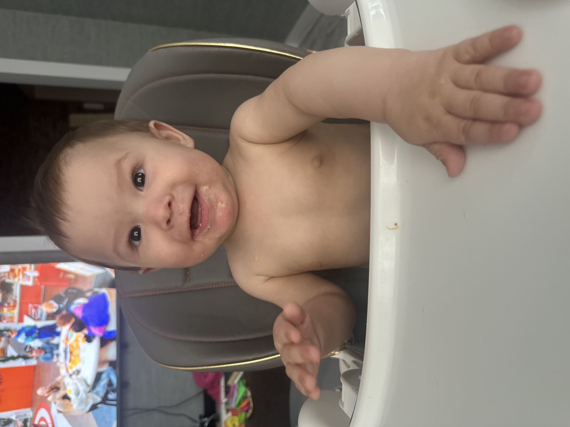 A toddler is sitting in a high chair, looking at the camera with a messy mouth. The toddler is shirtless and has food smeared on their face and chest. The high chair is white with a gray and gold seat. In the background, a television screen shows a group of people sitting around a table. The room appears to be a home environment.