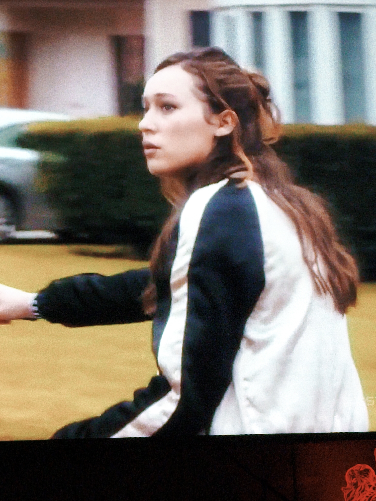 A medium shot shows a woman from the chest up, with her head turned to the right, looking away from the camera. She has long, dark brown hair pulled back into a ponytail. She is wearing a jacket with black sleeves and a white body, with a dark stripe running down the front. The background is blurred, but appears to be an outdoor setting with a lawn and a hedge. There is also a hint of a building and a vehicle in the background. The lighting is soft and natural. The image is grainy and appears to be a still from a video. There is no indication of the location being Vladivostok, Russia.